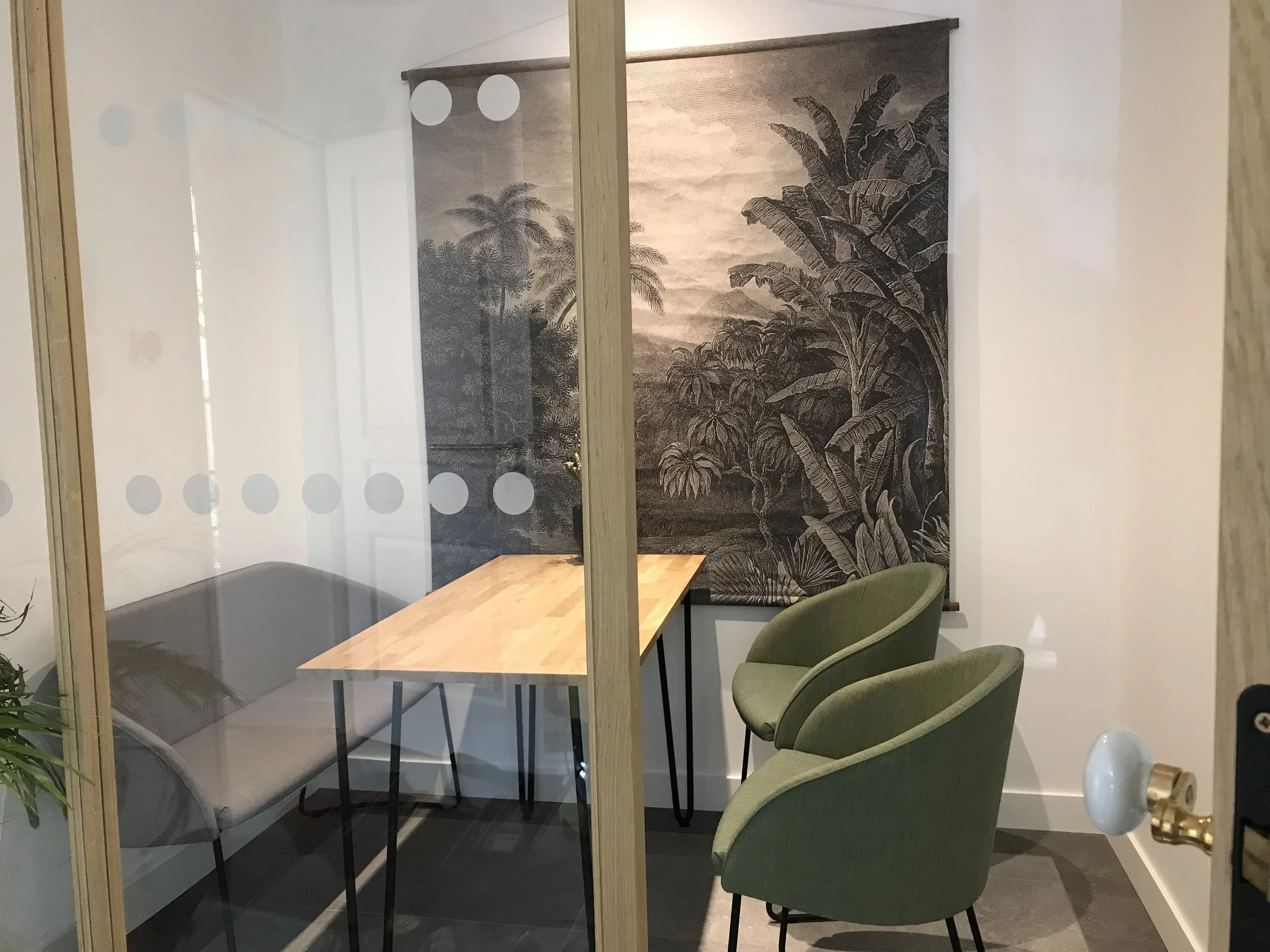 Salle de réunion avec un grand mur en arrière-plan décoré avec une toile d'un paysage tropical en noir et blanc. La pièce est séparée par une cloison en verre qui a des points circulaires. Il y a une table en bois avec des chaises vertes et grises autour.