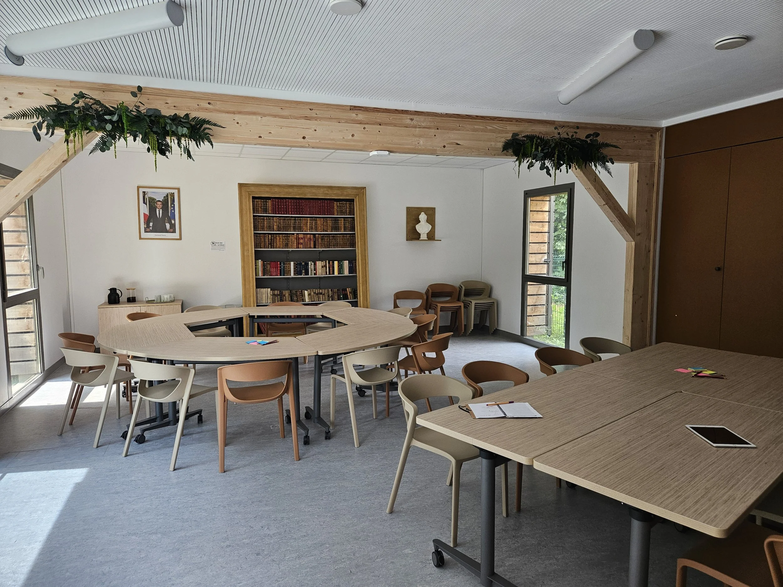 Salle de réunion avec tables en forme de U, fauteuils en bois, bibliothèque avec livres, décor avec statue blanche, fenêtres ouvertes laissant entrer la lumière naturelle, plantes vertes en suspension au plafond.