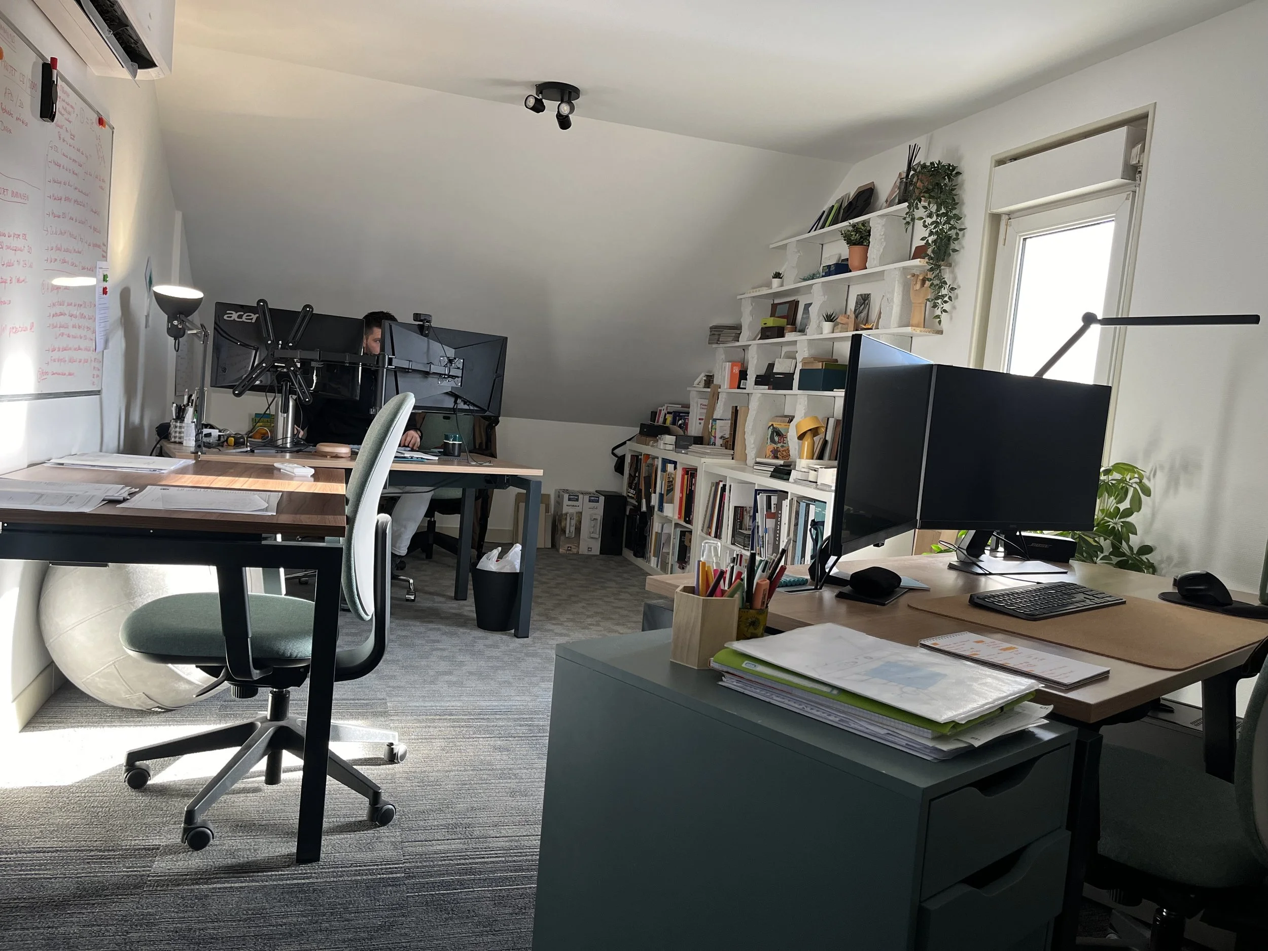 Bureau avec plusieurs bureaux, étagères remplies de livres et plantes, deux moniteurs d'ordinateur, une lampe, un tableau blanc avec écriture manuscrite, et des chaises. La pièce est bien éclairée par la lumière naturelle.