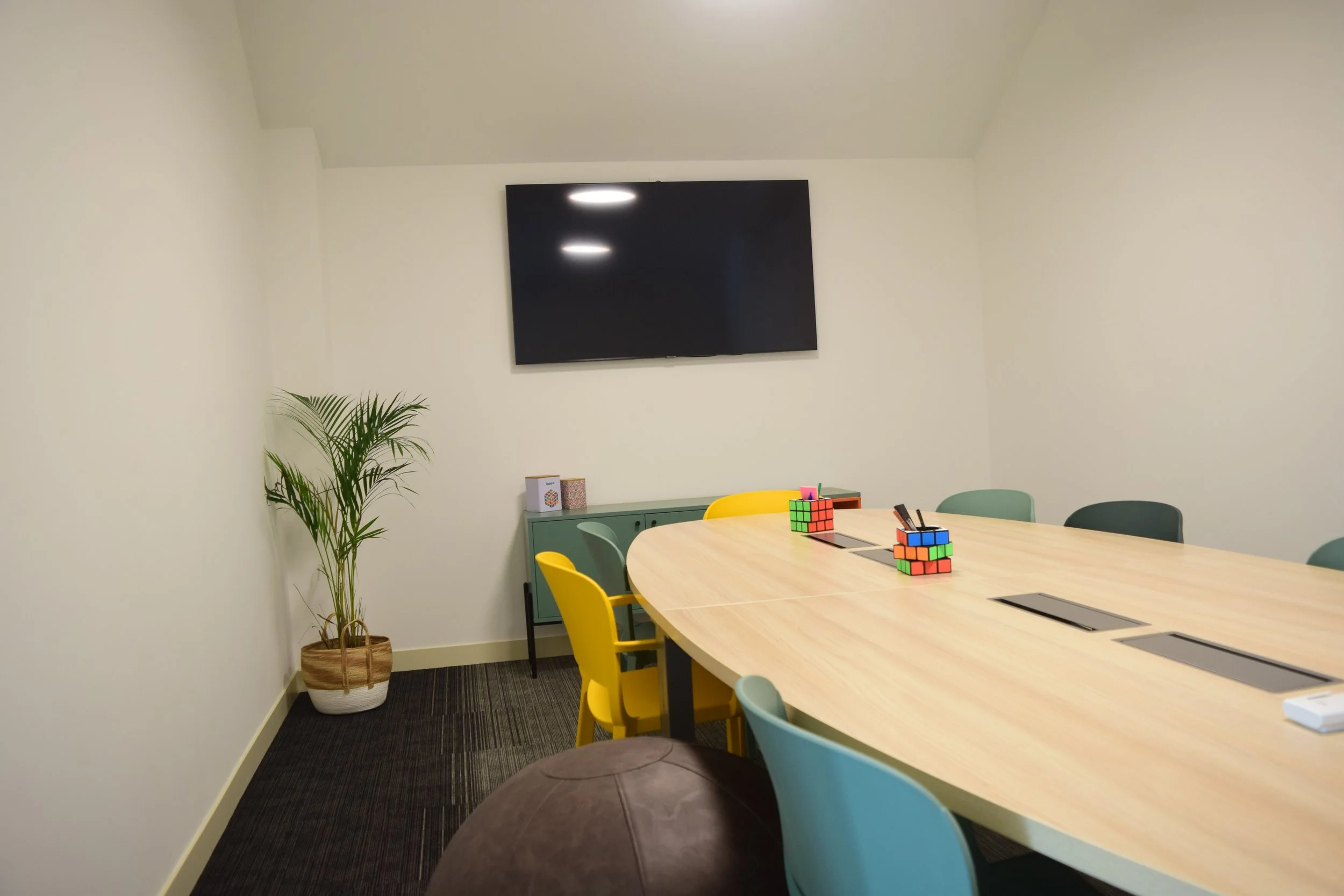 Salles de réunion avec un grand tableau écran, une table ovale en bois clair, des chaises de différentes couleurs, deux puzzles Rubik’s cubes, une plante verte et une bibliothèque en vert. Mur blanc, sol recouvert d’un tapis noir.