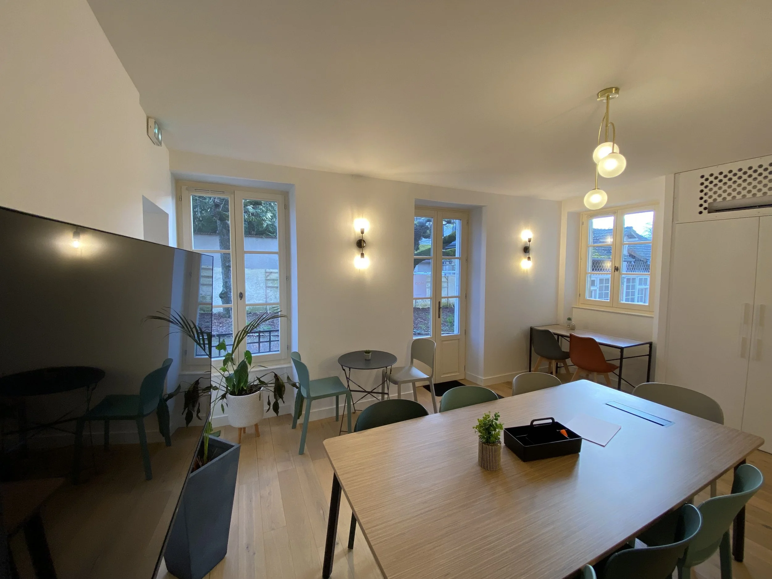 Salle de réunion lumineuse avec plusieurs fenêtres, mobilier moderne, plantes, et éclairage suspendu