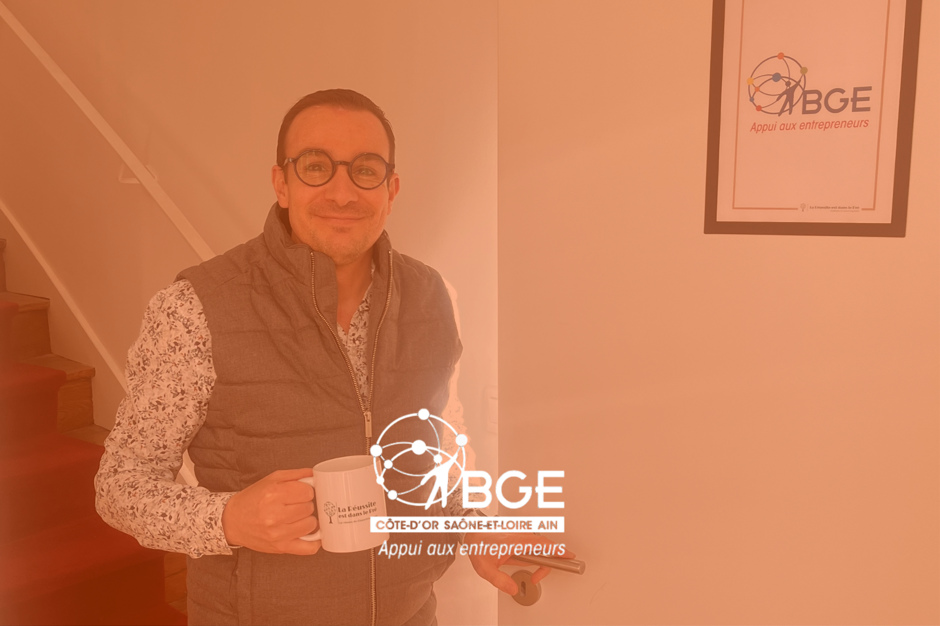 Un homme souriant portant des lunettes tient une tasse blanche dans une main et une poignée de canapé dans l'autre. Il se tient à côté d'un mur blanc où est affiché un panneau avec un logo de BGE, qui signifie 'Appui aux entrepreneurs', et le texte 'Côte-d'Or Saône-et-Loire Ain'.