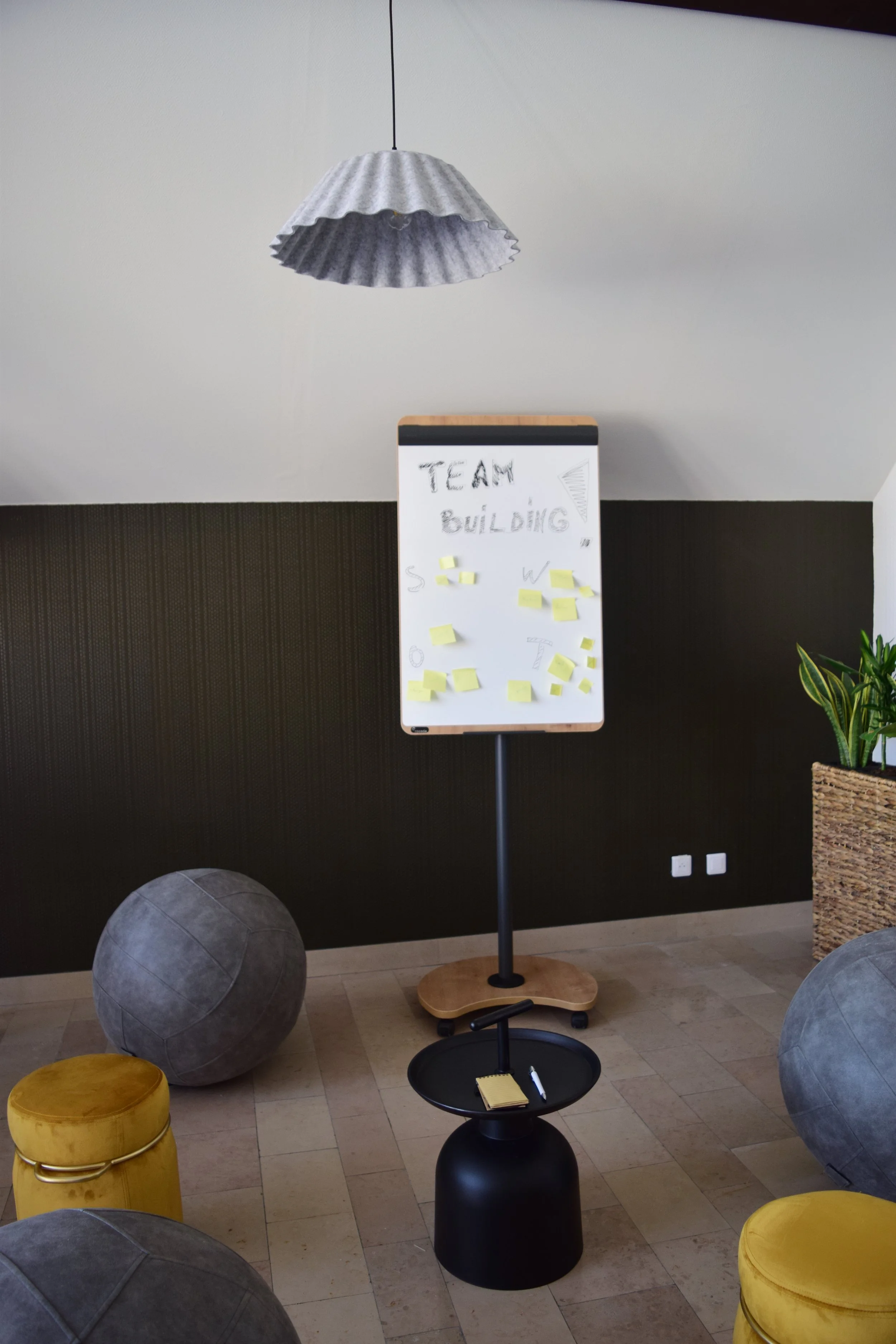 Salle de réunion avec tableau blanc, ballons de massage gris, pouf jaune, plante verte et lampadaire suspendu.