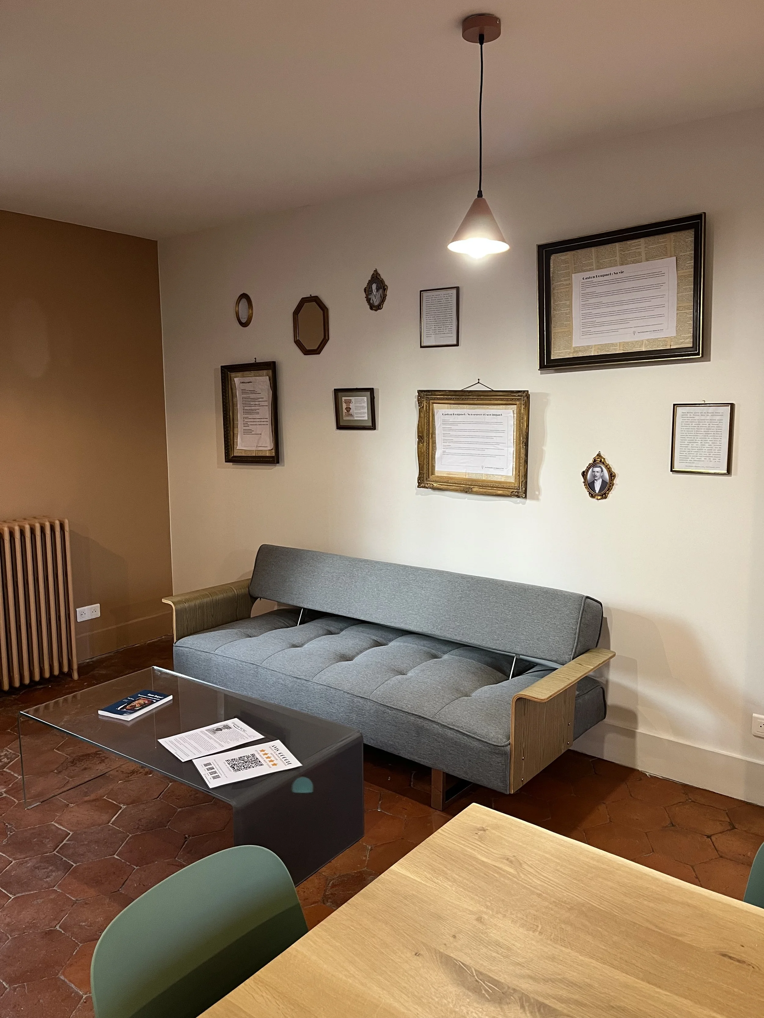 Salon intérieur avec un canapé gris, une table en verre, des œuvres d'art accrochées au mur et une lampe suspendue blanche. Il y a aussi un coin avec une table en bois et une chaise verte.