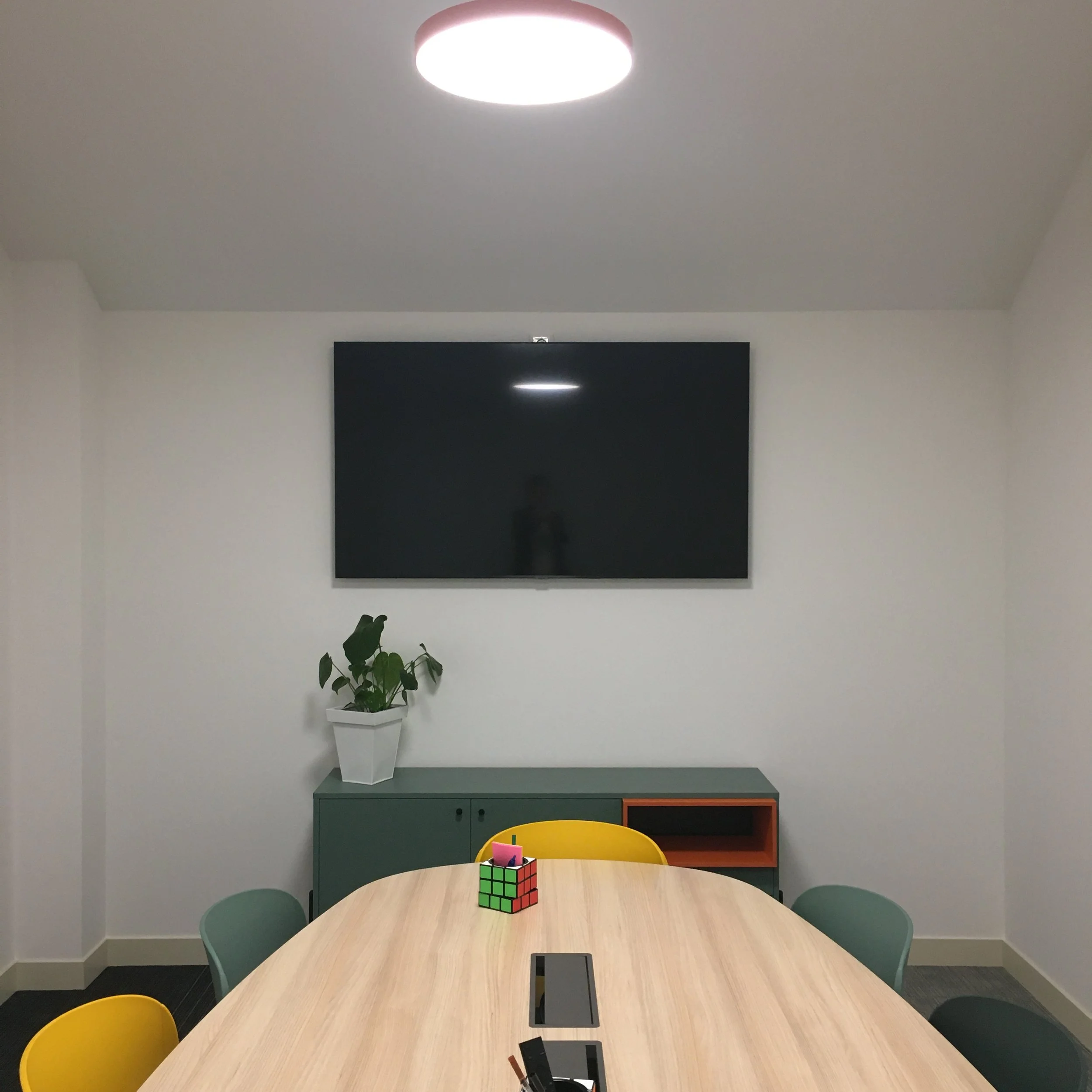 Salle de réunion avec table ovale, chaises vertes, une plante en pot blanche, un mur blanc avec une télévision noire, un plafond avec une lumière ronde, un Rubik's Cube sur la table, et un bouton d'extinction à proximité.