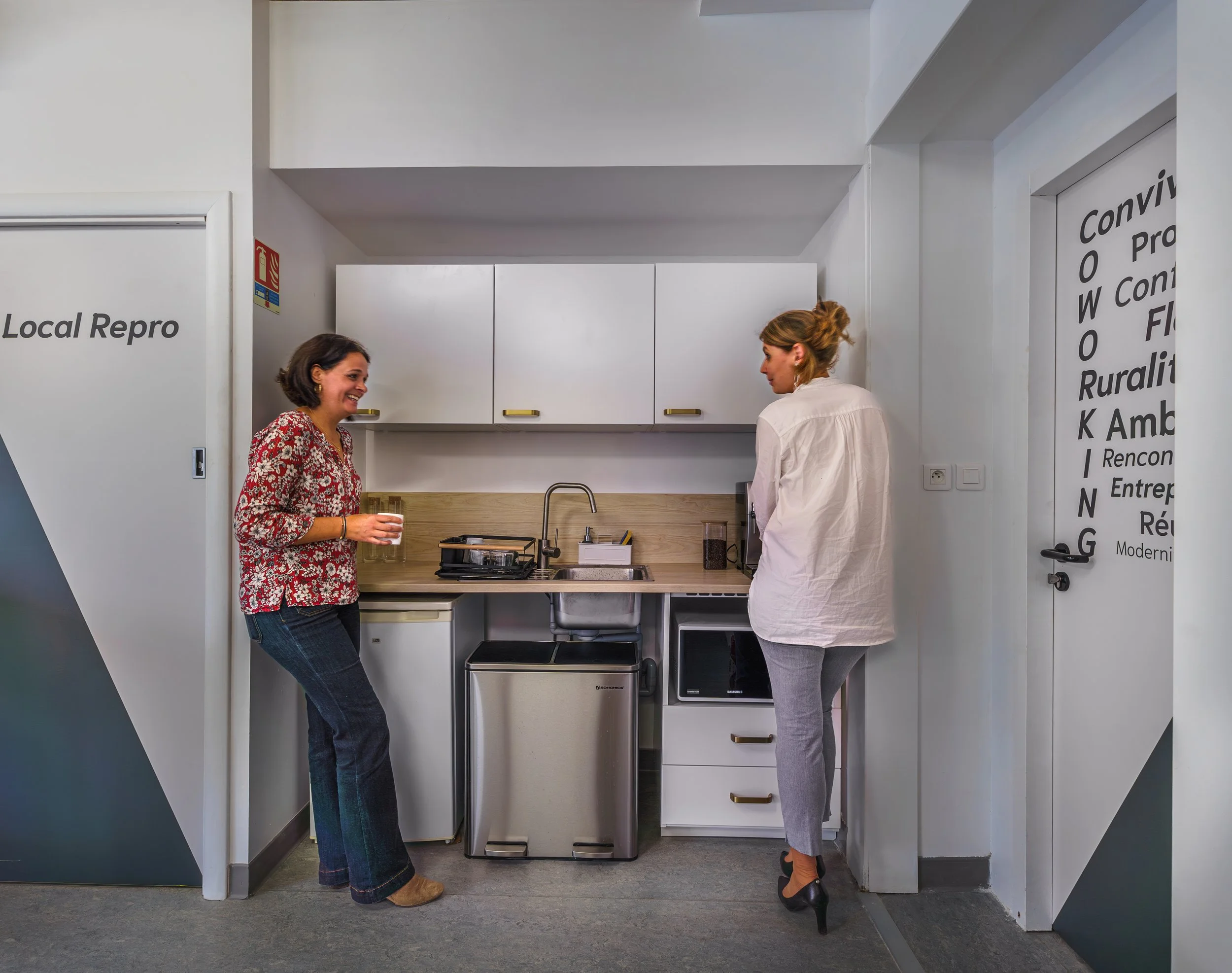 Deux femmes discutent dans une cuisine moderne, l'une porte un chemisier à fleurs rouge, l'autre un chemisier blanc, elles semblent souriantes et engagées dans leur conversation.