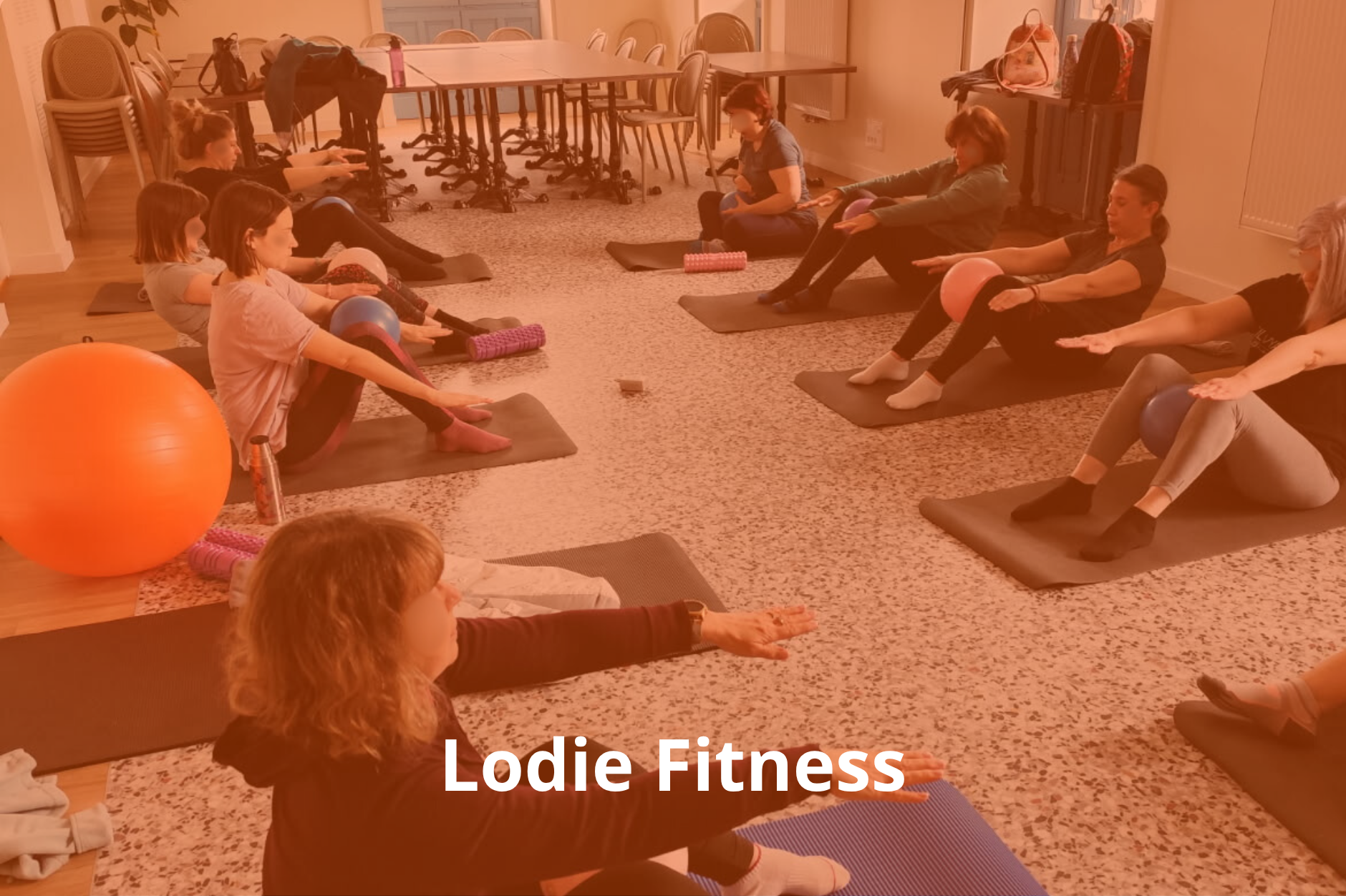 Groupe de personnes, principalement des femmes, pratiquant le yoga ou une activité de fitness sur des tapis, dans une salle intérieure. Certaines utilisent des ballons, et il y a un grand ballon orange et divers accessoires de sport autour.