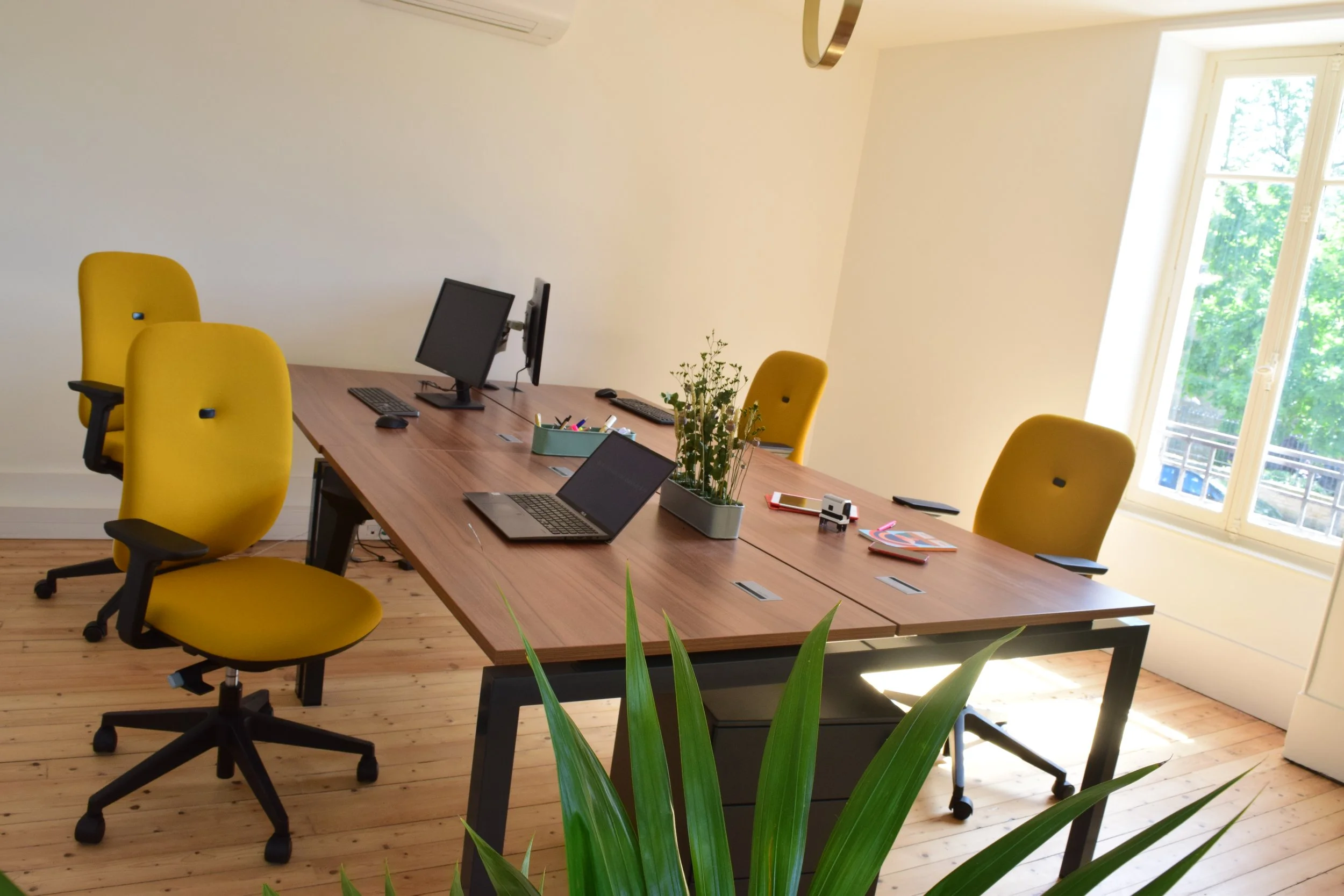 Une salle de réunion moderne avec une grande table en bois, entourée de chaises jaunes. Sur la table, il y a deux ordinateurs portables, des plantes, des stylos et des papiers. La pièce est bien éclairée par une grande fenêtre laissant entrer la lumière naturelle et offrant une vue sur des arbres verdoyants.