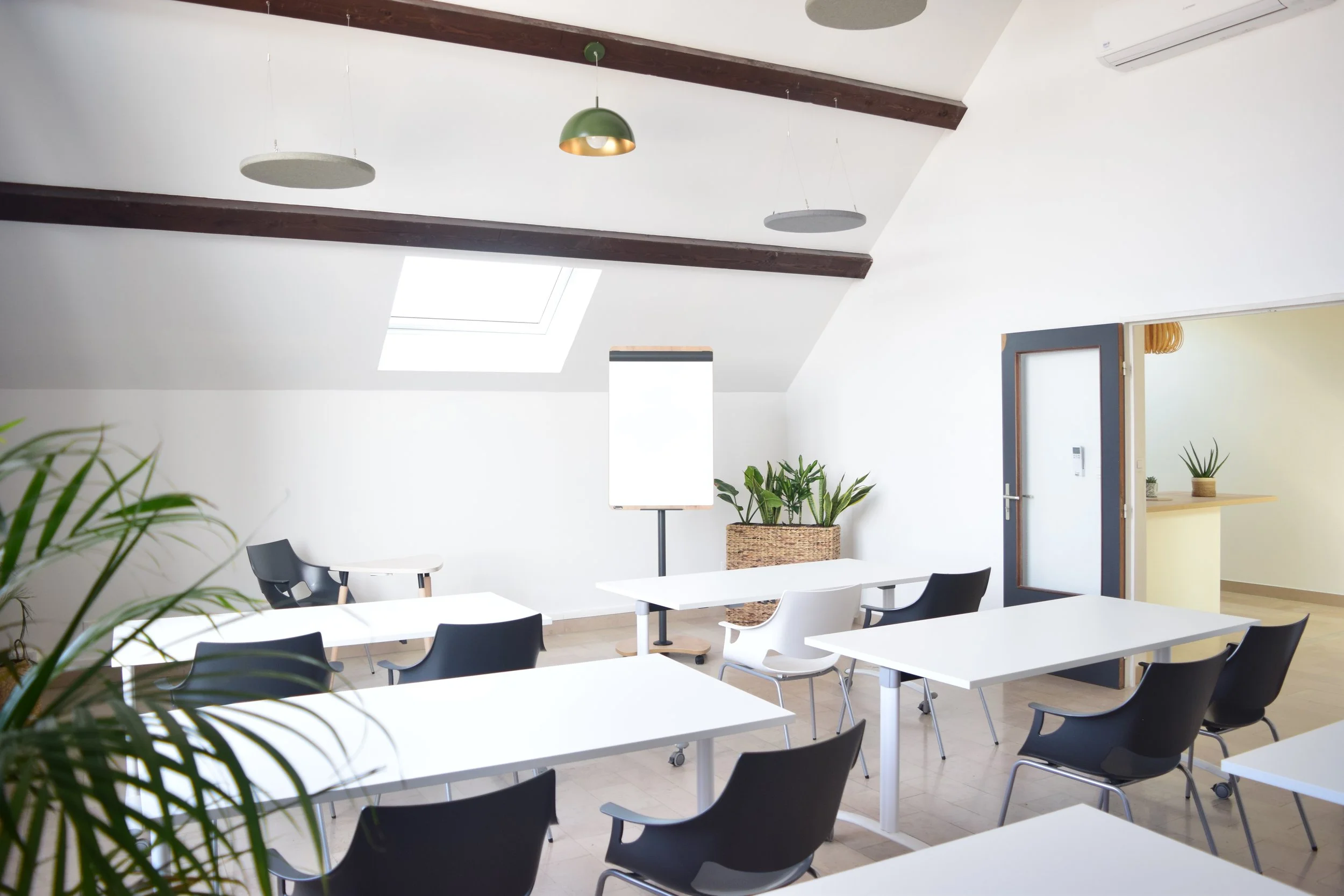 Salle de réunion ou de formation avec des tables blanches, des chaises noires, des plantes vertes, un éclairage suspendu et une fenêtre de toit, en intérieur moderne
