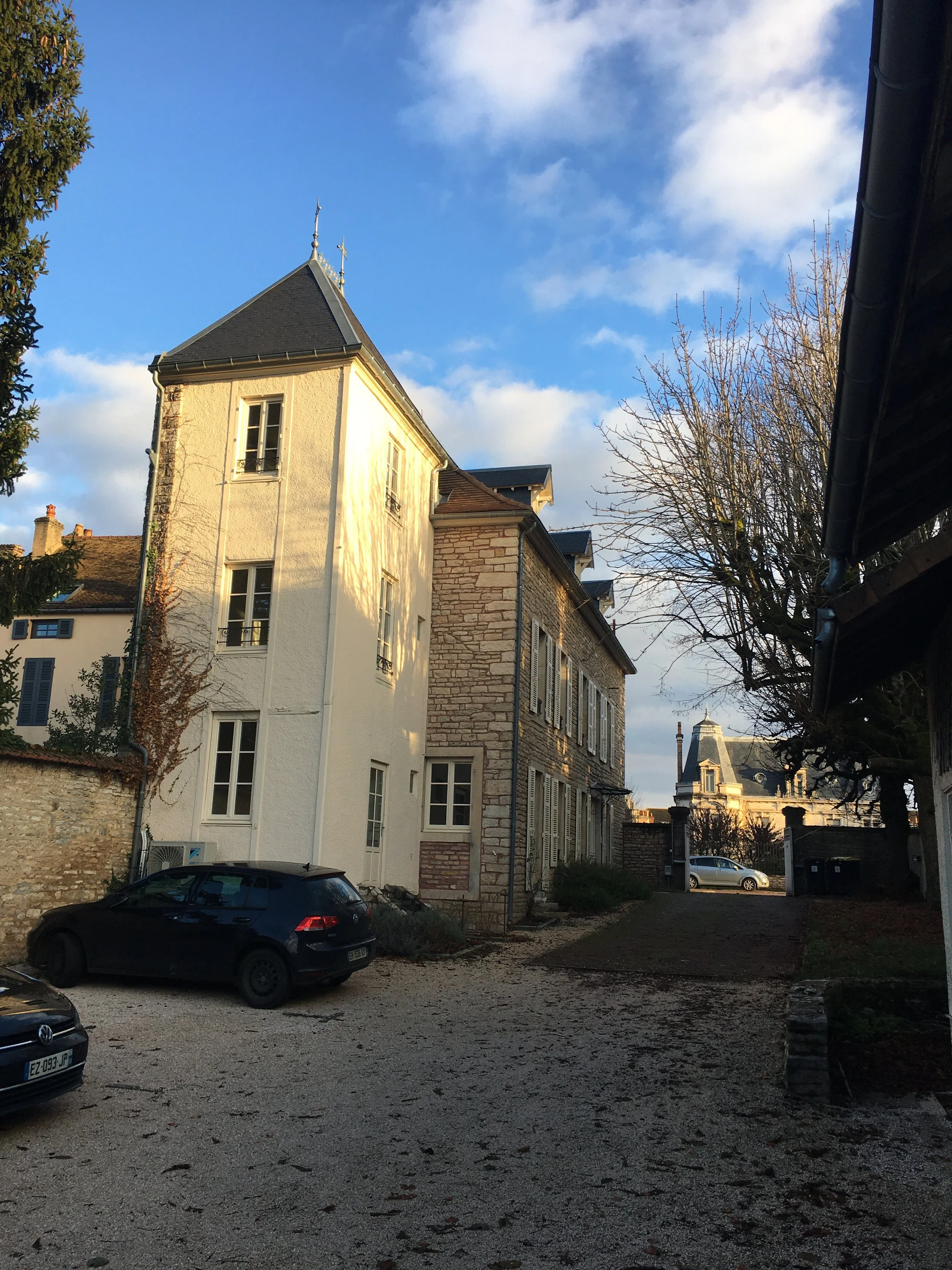 Une cour pavée avec des voitures garées, entourée de bâtiments en pierre et en plâtre, avec des arbres dénudés sous un ciel en partie nuageux.