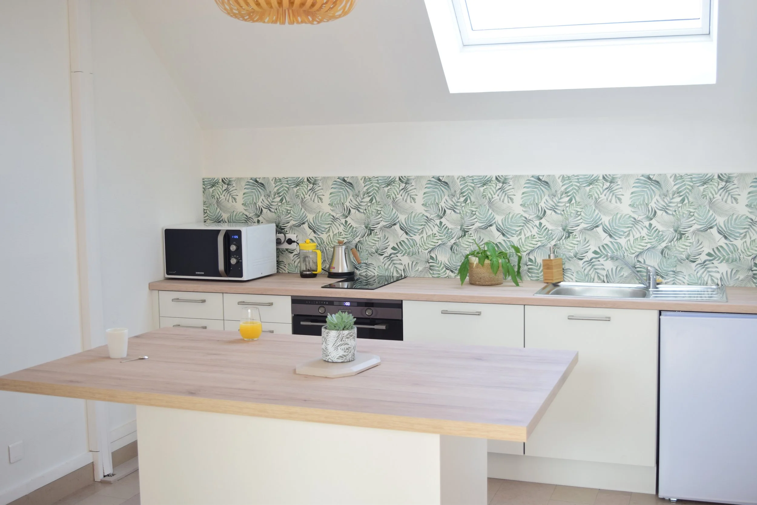 Cuisine moderne avec un plan de travail en bois clair, un micro-ondes noir et blanc, une carafe d'eau, un pot de plantes vertes, un espoir électrique, un évier avec un robinet, un buffet blanc et un mur avec un papier peint à motifs de feuilles vertes. Un plafond avec un velum en bois orangé et une lucarne au plafond.