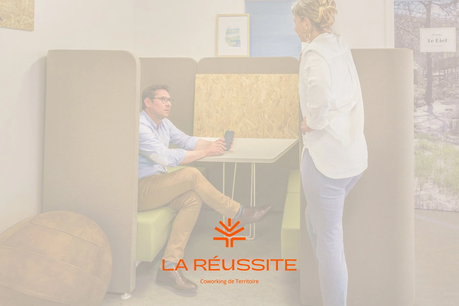 Deux personnes discutent dans un espace de coworking, une homme assis utilisant son téléphone et une femme debout face à lui, avec un mur blanc, des tableaux et une fenêtre en arrière-plan, texte superposé : 'LA RÉUSSITE - Coworking de Territoire'.