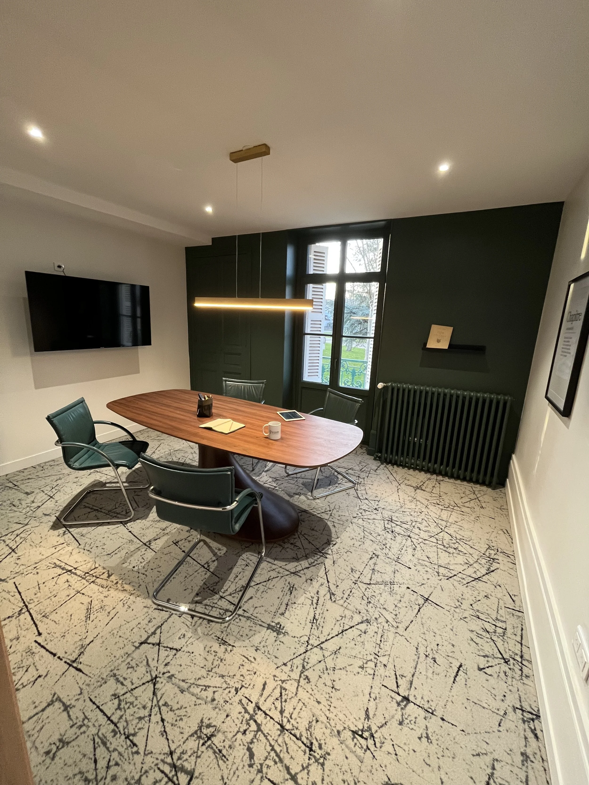 Salle de réunion avec table en bois, cinq chaises vertes avec bras en métal, un mur avec une télévision murale, une fenêtre avec volets en bois, et un mur vert foncé avec une étagère et un cadre.