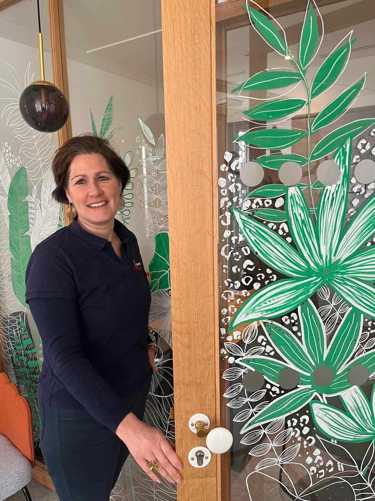 Une femme souriante se tient à côté d'une porte en bois avec des décorations murales de plantes vertes. Elle porte un pull noir et des boucles d'oreilles géométriques. La porte est ornée d'illustrations de plantes vertes stylisées.