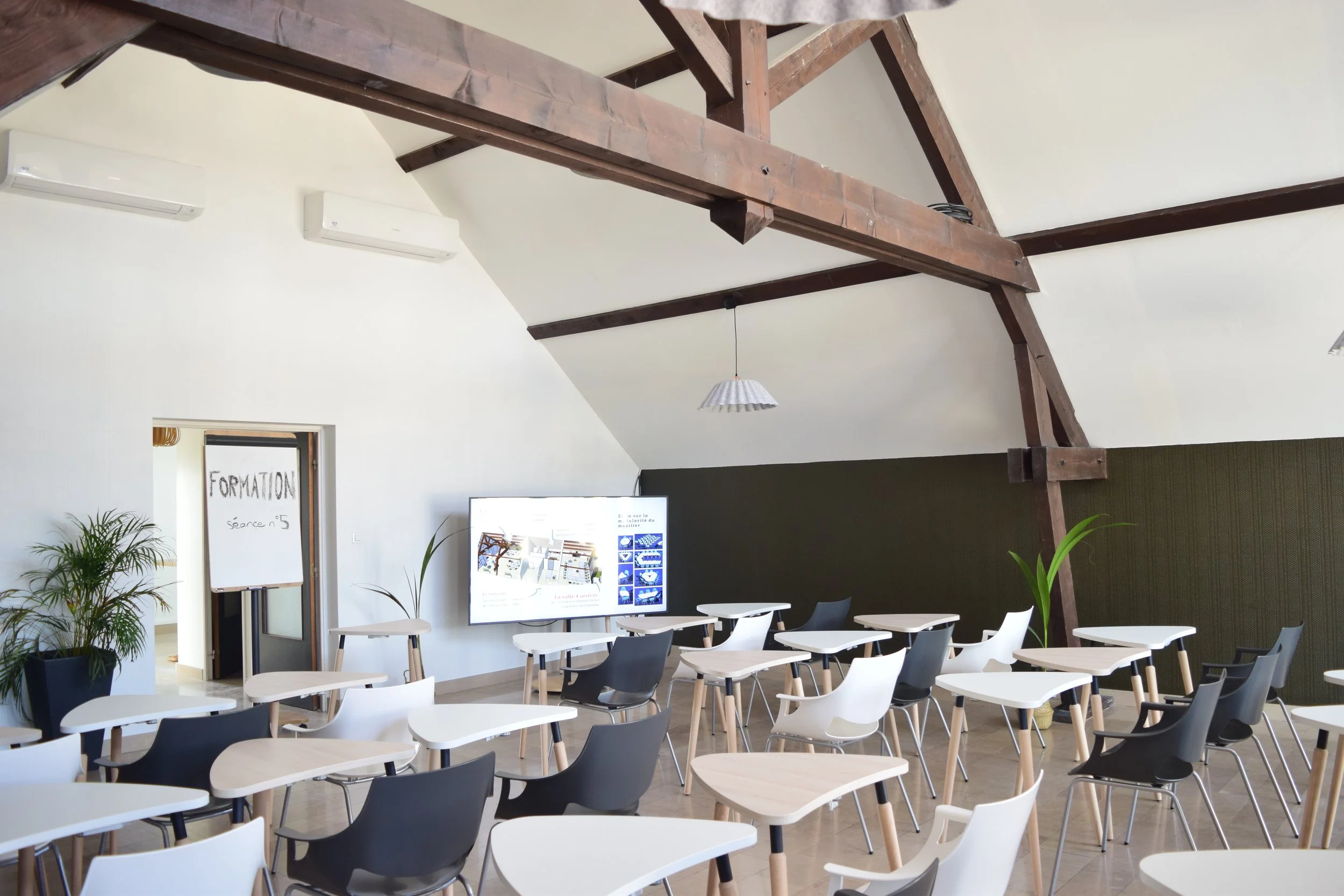Salle de réunion avec des chaises blanches et noires, une grande télévision, des plantes en pot, un tableau blanc avec l'inscription "FORMATION" et un plafond avec poutres en bois.