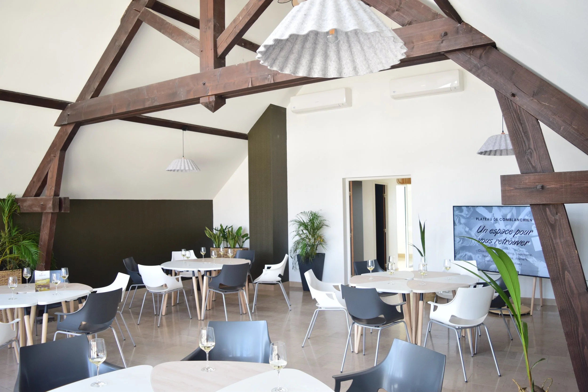 Salle à manger élégante avec des tables rondes habillées de nappes blanches et des verres de vin, décorée de plantes vertes et éclairée par des lampes suspendues, avec une structure en bois au plafond.