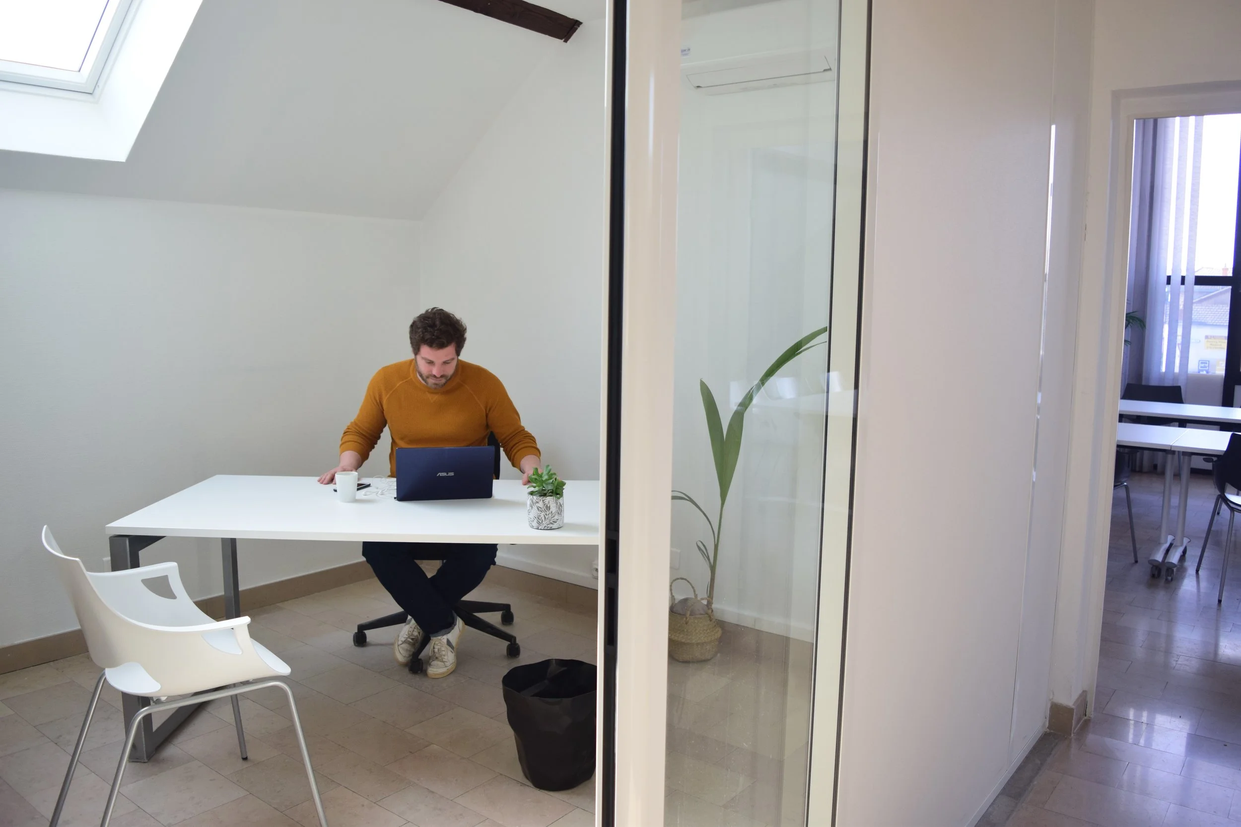 Un homme assis à une table de bureau dans une salle avec plafond en pente et lumière naturelle provenant d'une fenêtre de toit. Il travaille sur un ordinateur portable, avec des tasses et une plante sur la table. La pièce est séparée par une cloison en verre.
