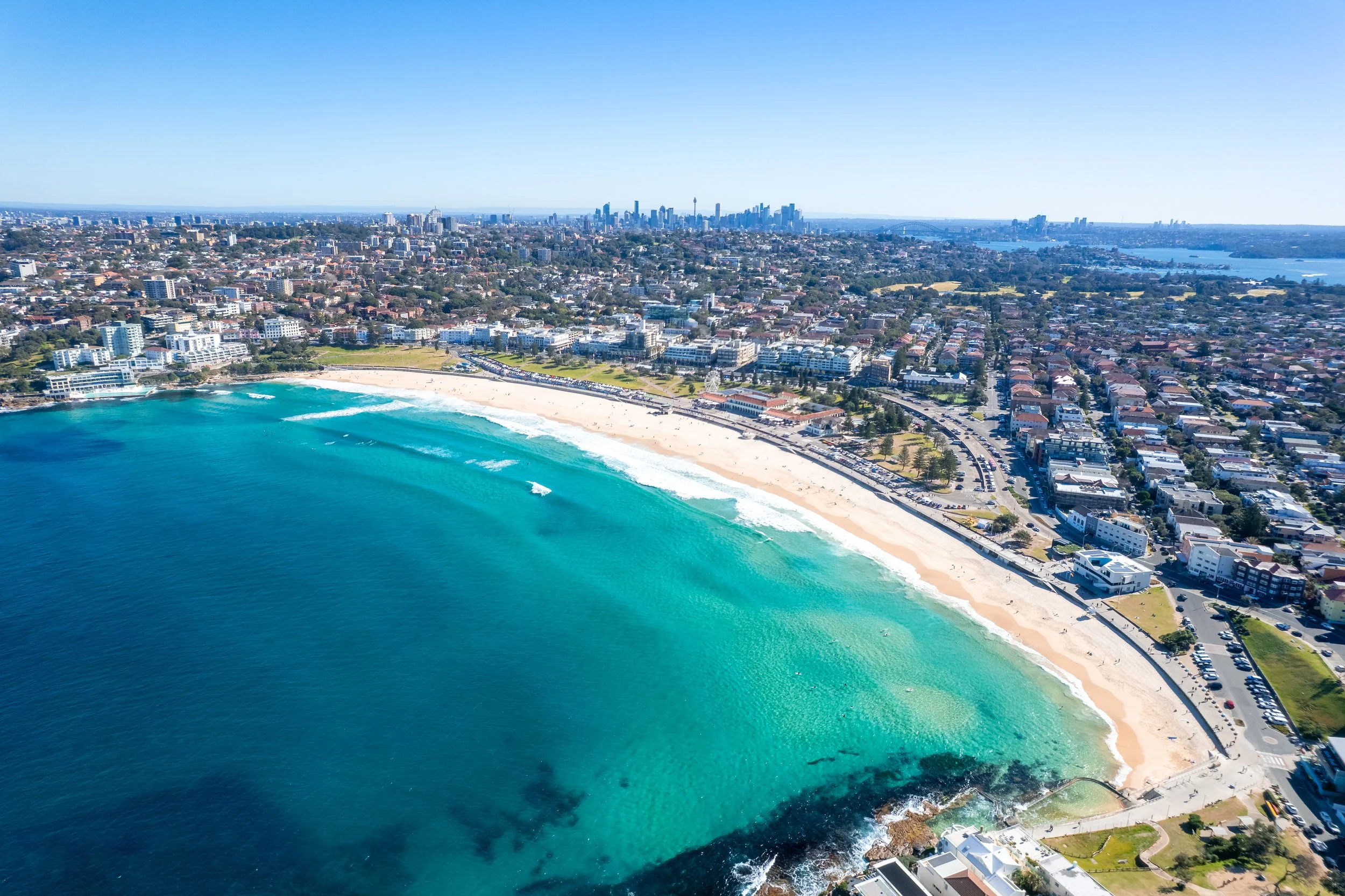 Aerial view of a sandy beach with turquoise water, city skyline in the background, and urban area alongside the coast.