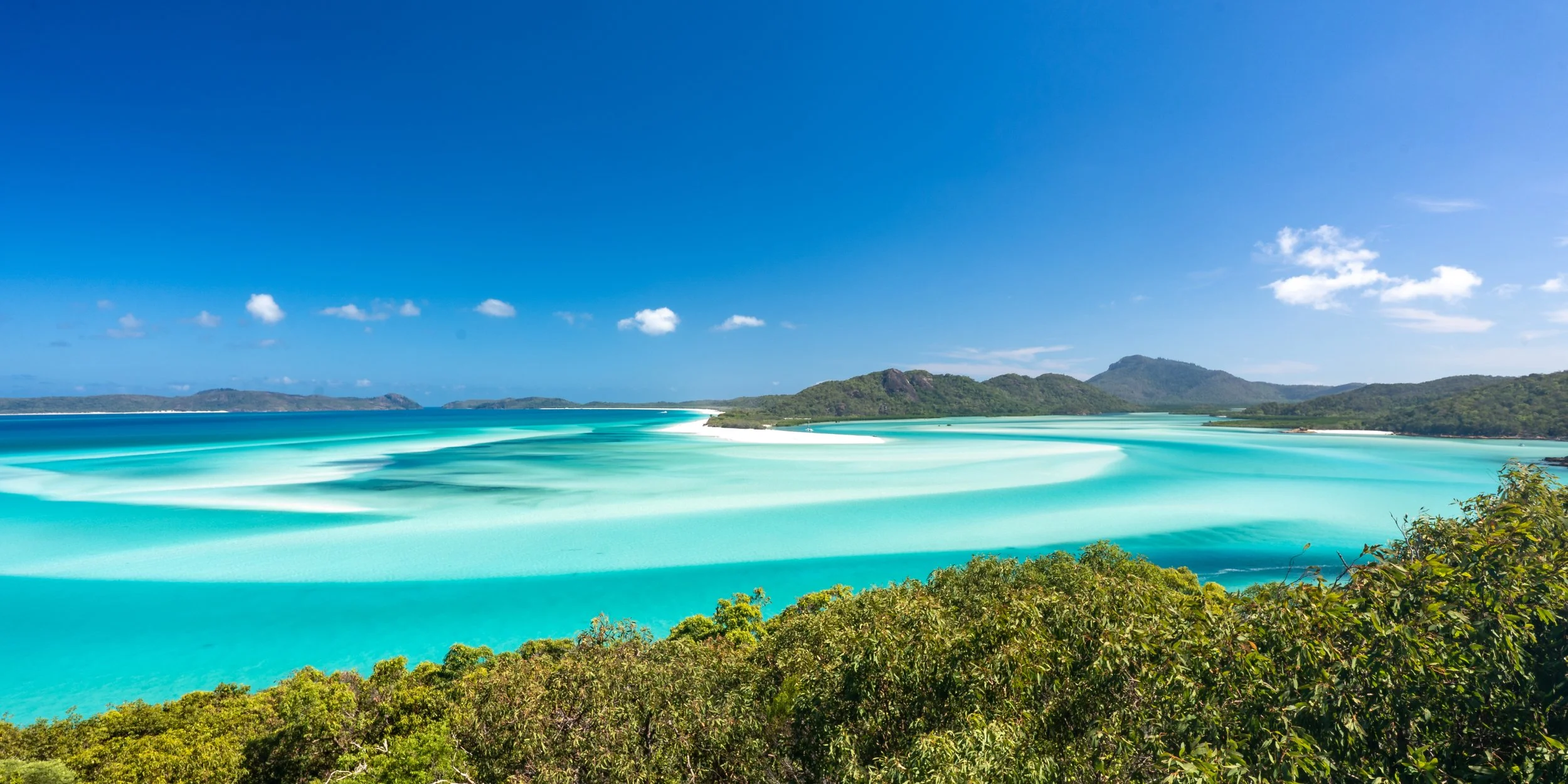 Scenic view of a coast with turquoise ocean, beach sandbars, lush greenery in the foreground, and hills and islands under a blue sky with clouds.