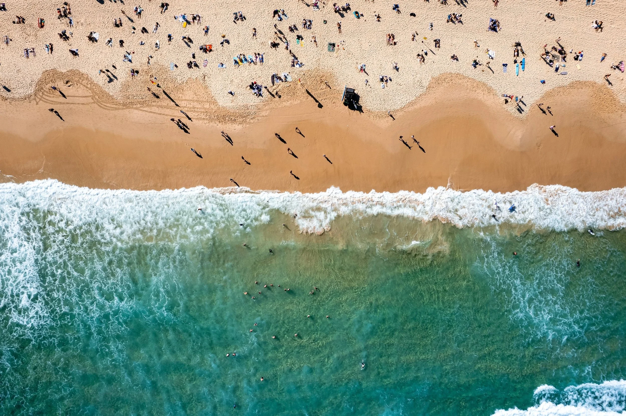 An aerial view of a beach showing people on the sandy shore and in the water, with waves crashing onto the sand.