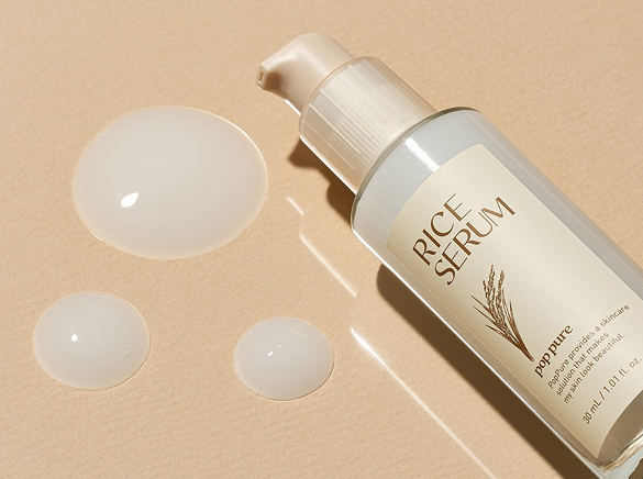 A bottle of rice serum with three drops of serum on a beige surface.