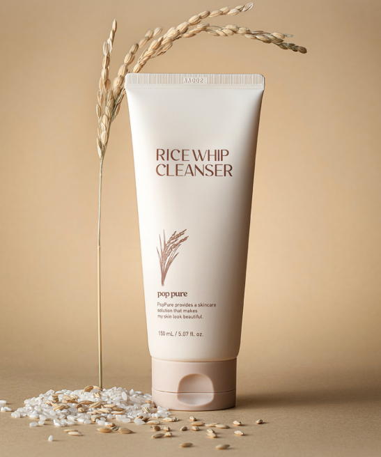 A tube of Rice Whip Cleanser labeled 'pop pure' stands upright on a beige surface with scattered oats and a stalk of oat grains behind it.