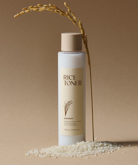 A bottle of rice toner with a beige cap, a rice stalk, and rice grains scattered on a neutral background.
