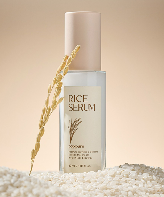 A bottle of Rice Serum with a beige pump cap, surrounded by rice grains and a rice stalk, against a neutral background.