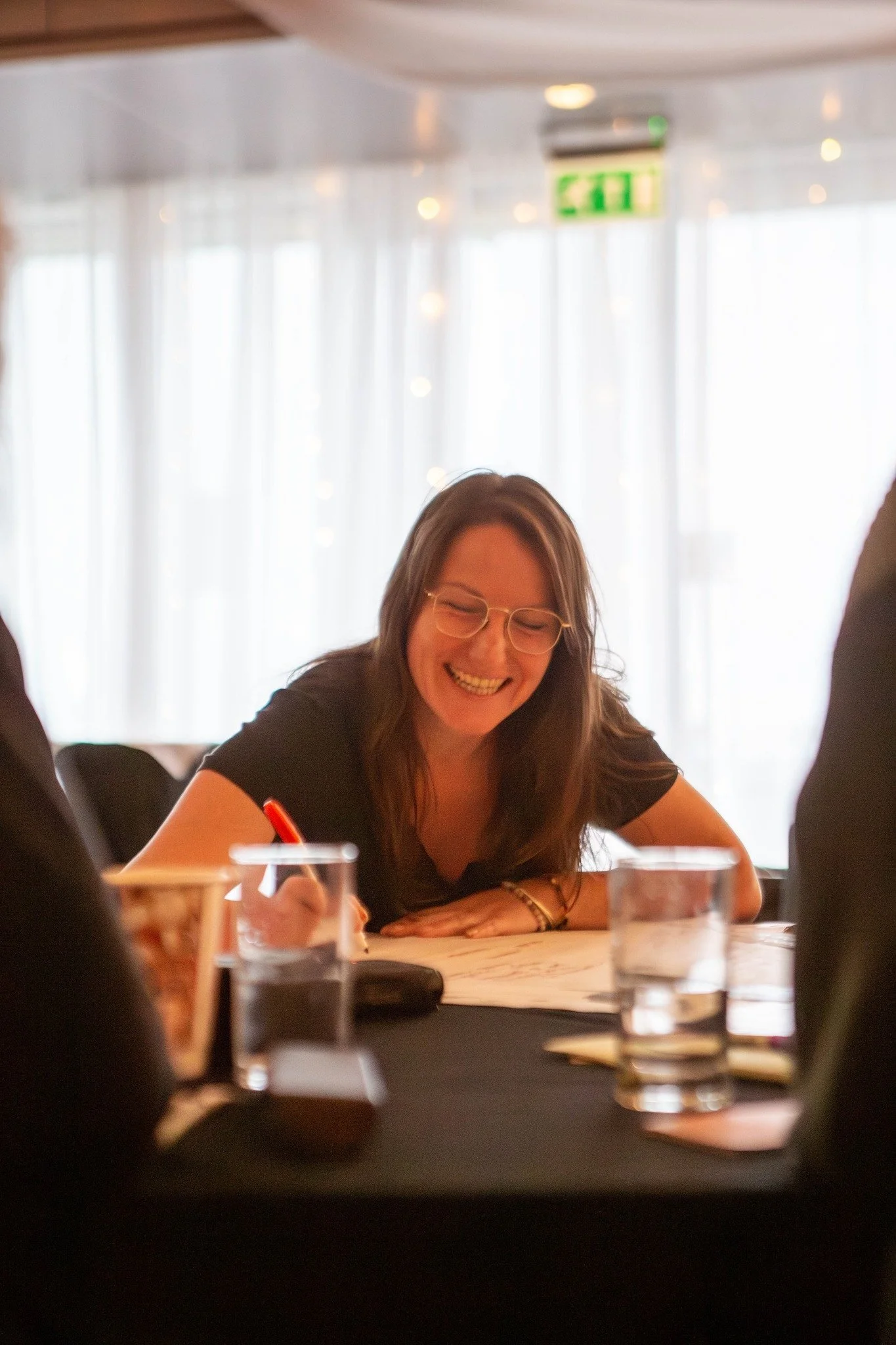 A woman with glasses smiling while sitting at a table with papers, glasses, and a pen in front of her in a bright room with sheer curtains and an exit sign in the background.