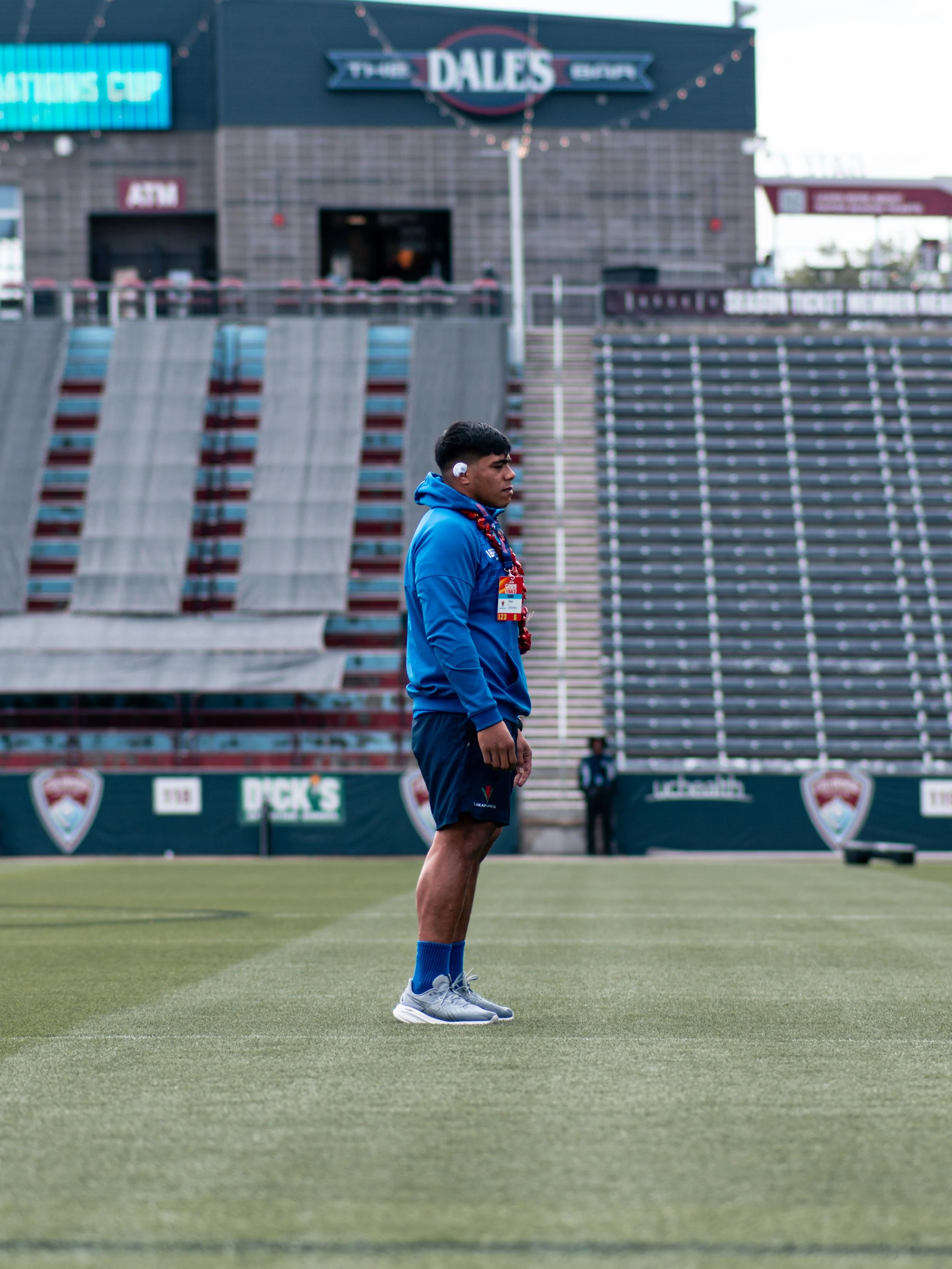 A young man in a blue sports outfit standing on a sports field with his hands on his hips, near stadium bleachers and event banners in the background.