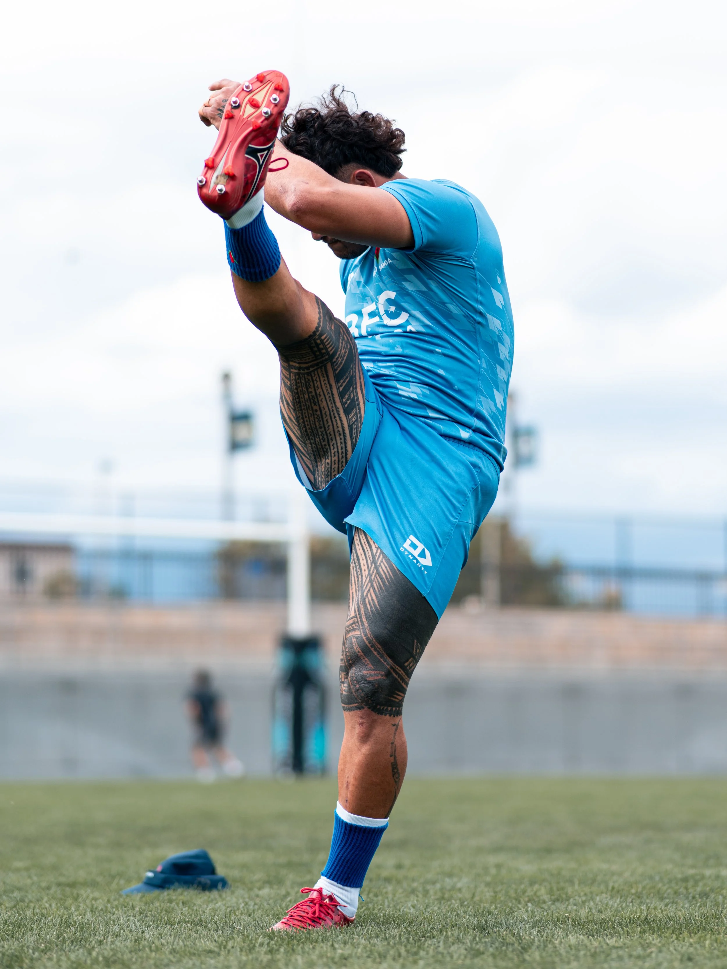 A male rugby player in a blue uniform stretching on a soccer field, holding his foot up behind him to stretch his quadriceps, with a cloudy sky in the background.