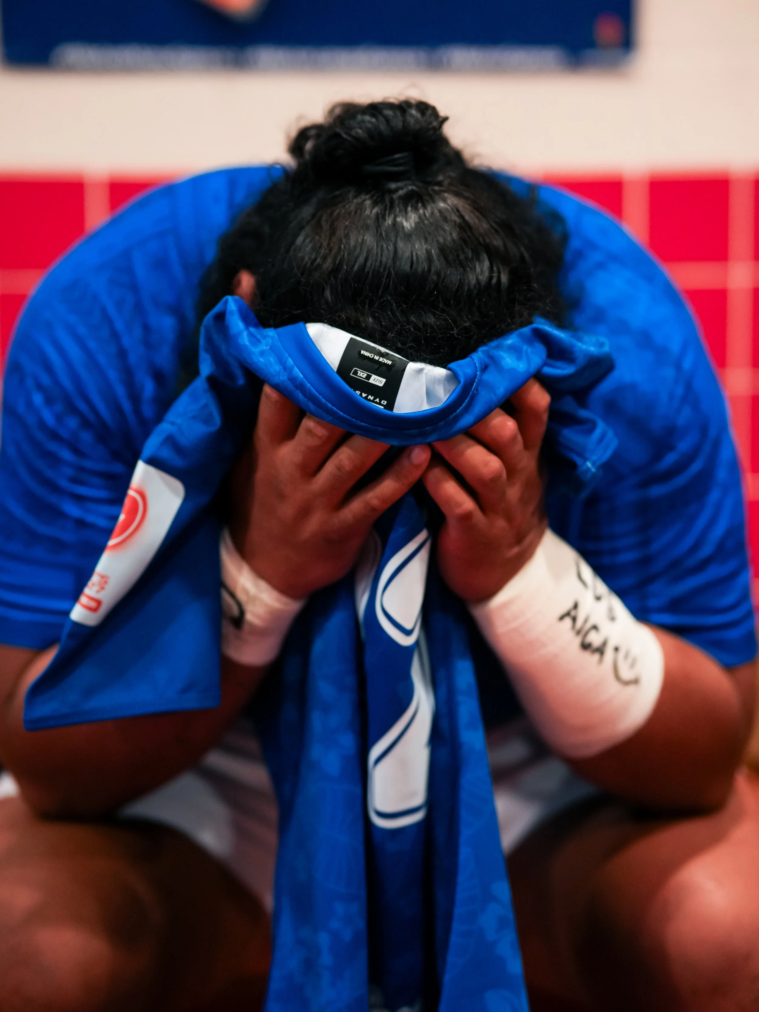 A person in blue sportswear covering their face with their hands, with a jersey on their head, in a sports environment.