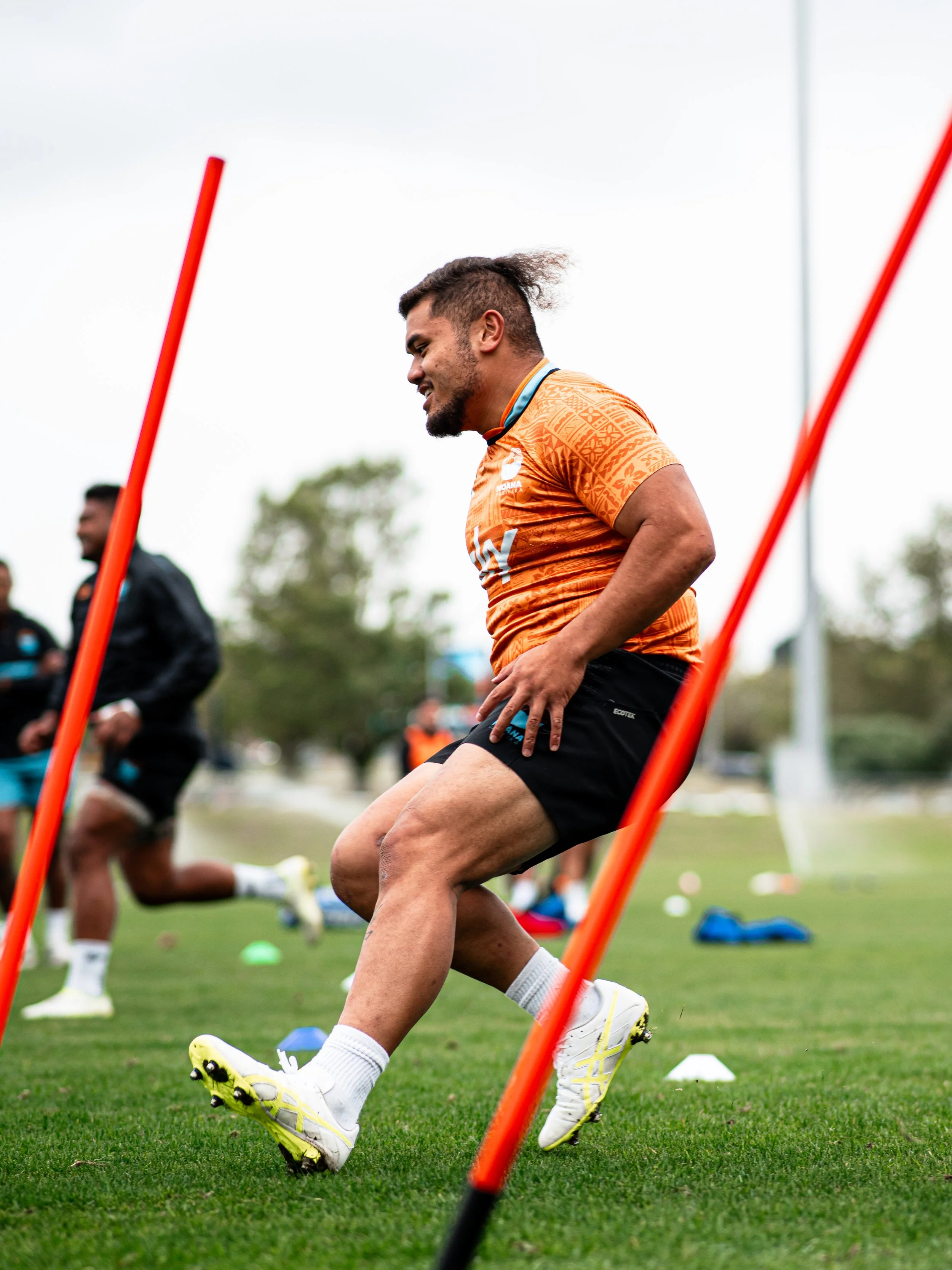Rugby player in orange jersey training on a green field, maneuvering around orange poles during practice.