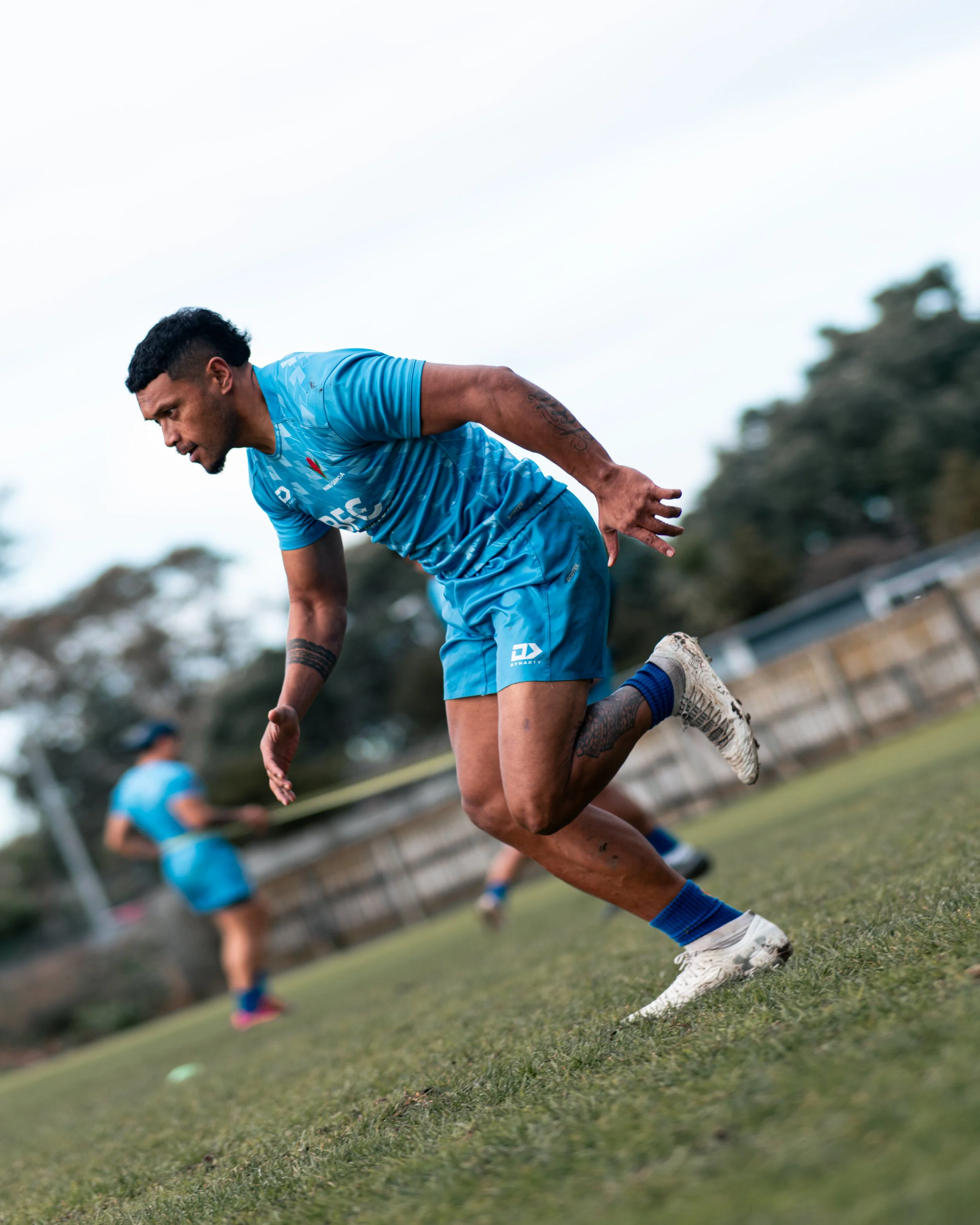 A male rugby player in a blue uniform is bending over and balancing on one leg on a grassy field during a game, with other players blurred in the background.