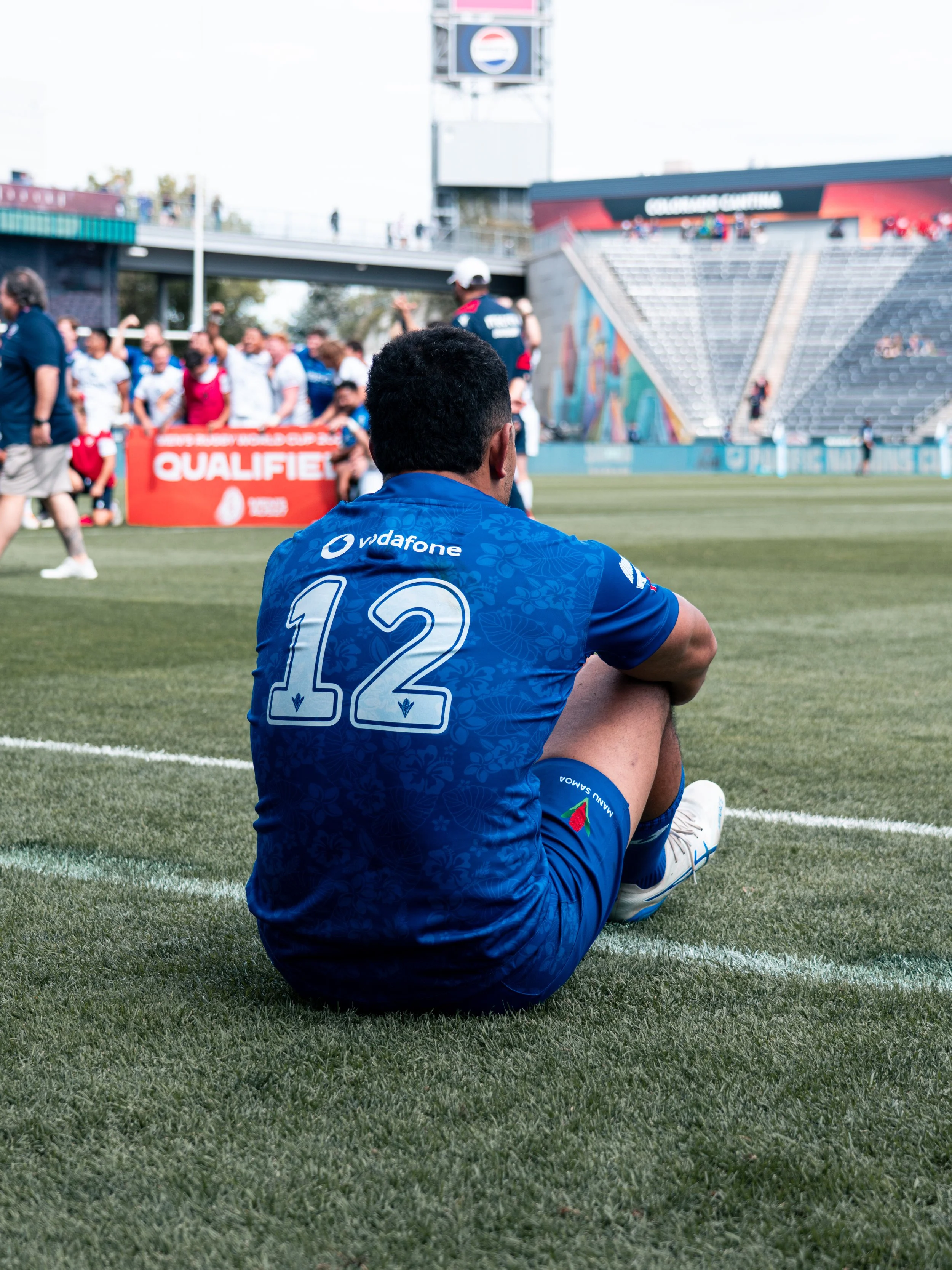 A rugby player sitting on the grass field, wearing a blue jersey with the number 12, with his back to the camera. In the background, a group of people and a few empty stadium seats are visible, along with a scoreboard and flags.