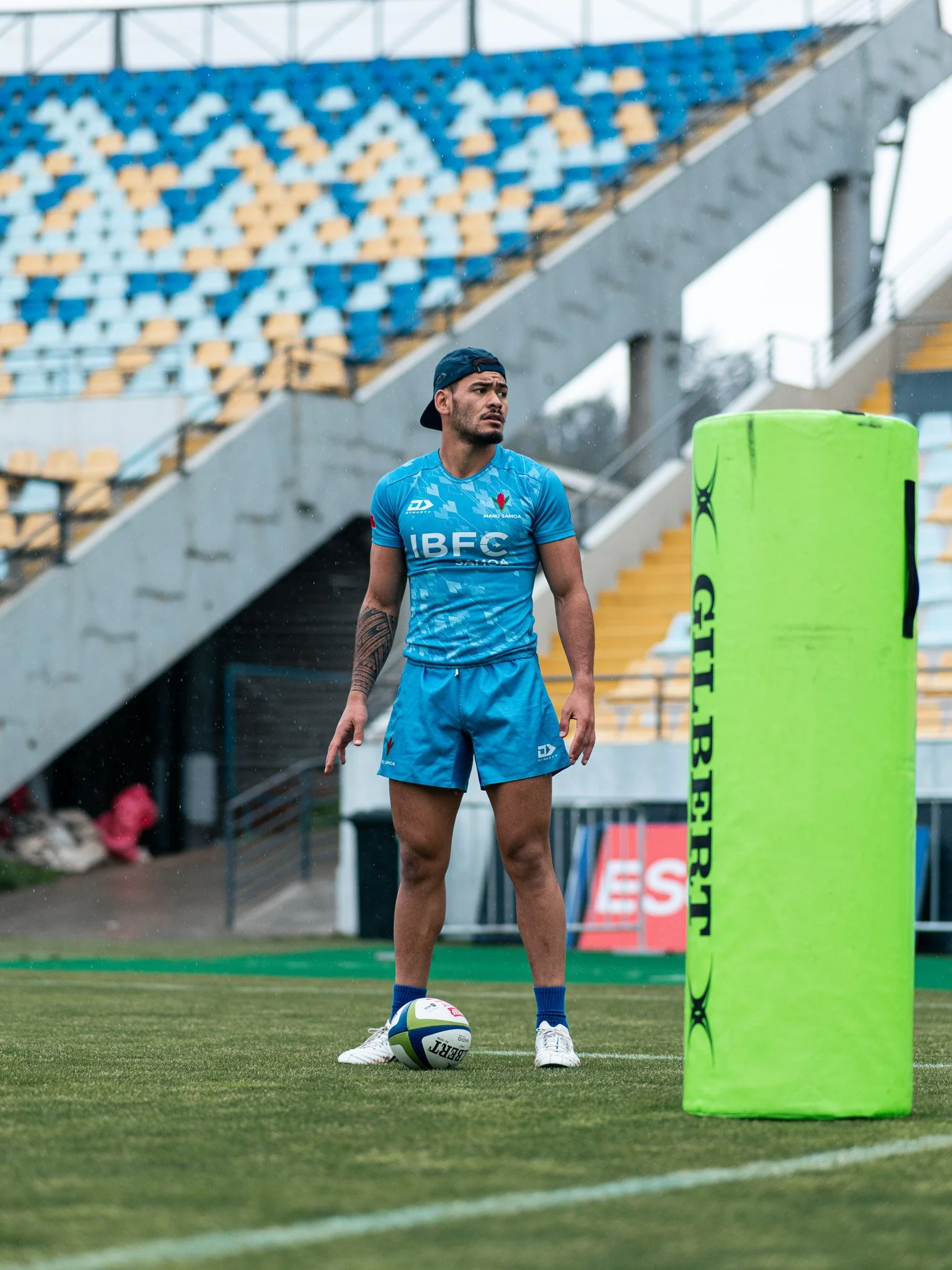 A rugby player standing on a field near a green tackle pad, with a rugby ball at his feet, wearing a blue uniform and a backwards cap, with empty stadium seats in the background.