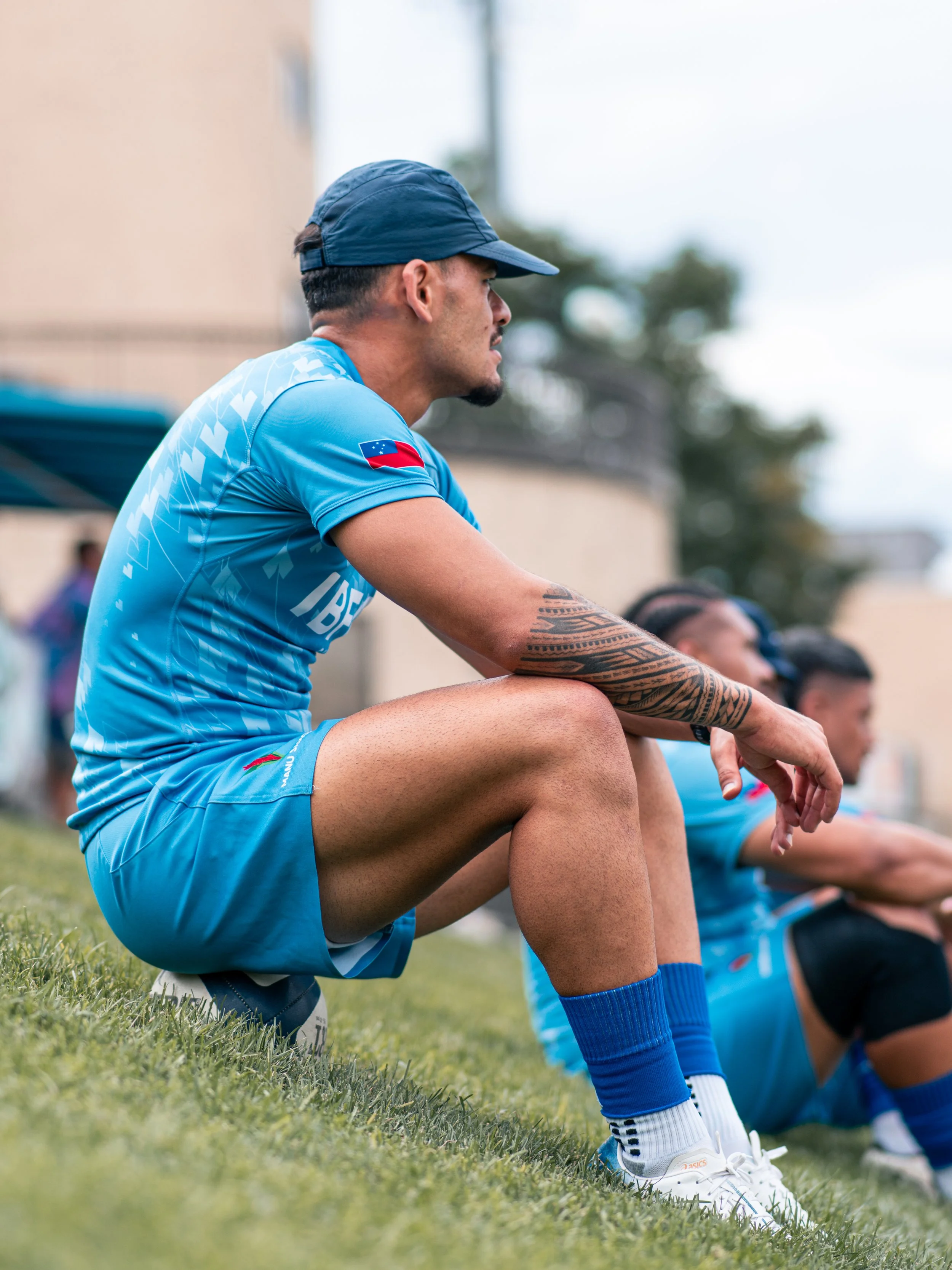 A group of athletes sitting on a grassy field during a sports practice, wearing blue uniforms and kneeling with arms resting on their knees.