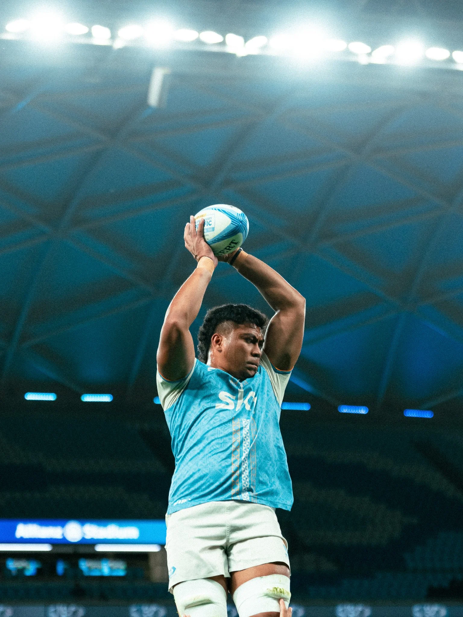 A rugby player in a light blue jersey preparing to throw the ball during a game in a stadium.