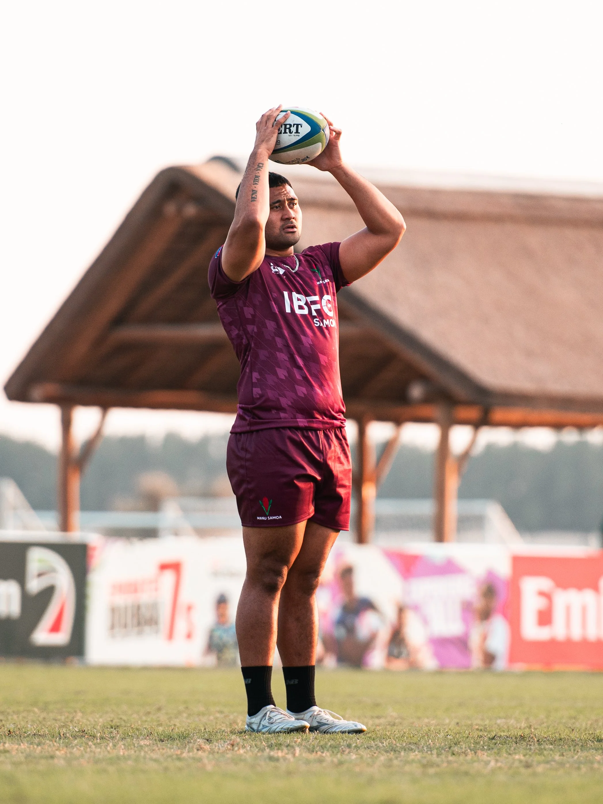A rugby player preparing to kick a rugby ball on the field, wearing a maroon uniform with the logos of Manu Samoa and IBFC. The background features a wooden structure and colorful advertising boards.