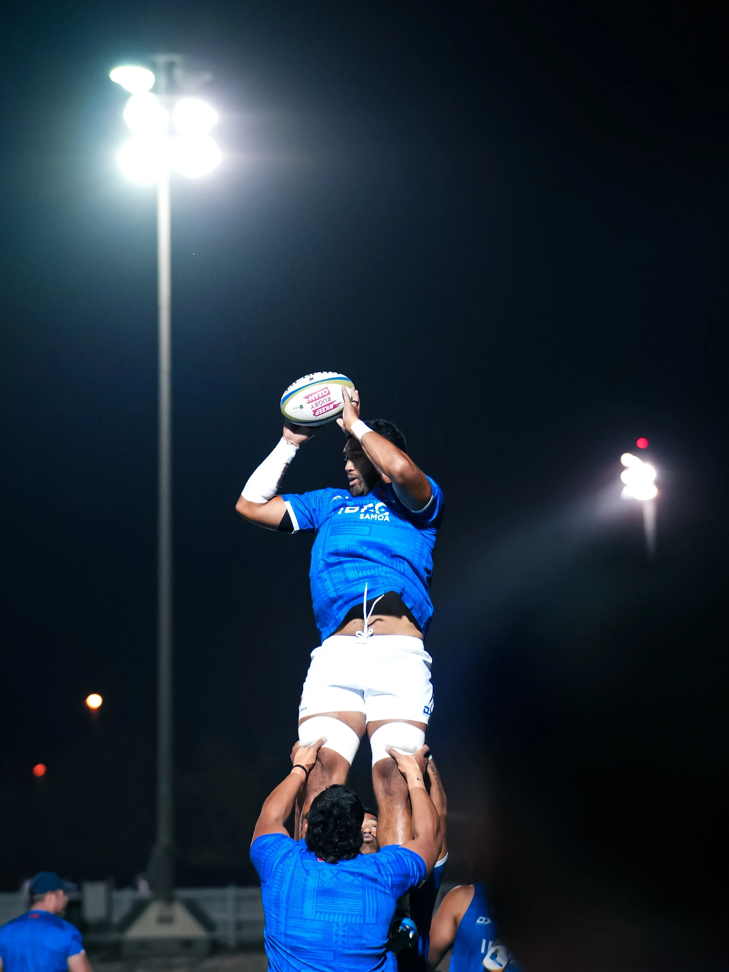 A rugby player in a blue uniform catches a rugby ball while being lifted by teammates during a night game, with stadium lights illuminating the scene.