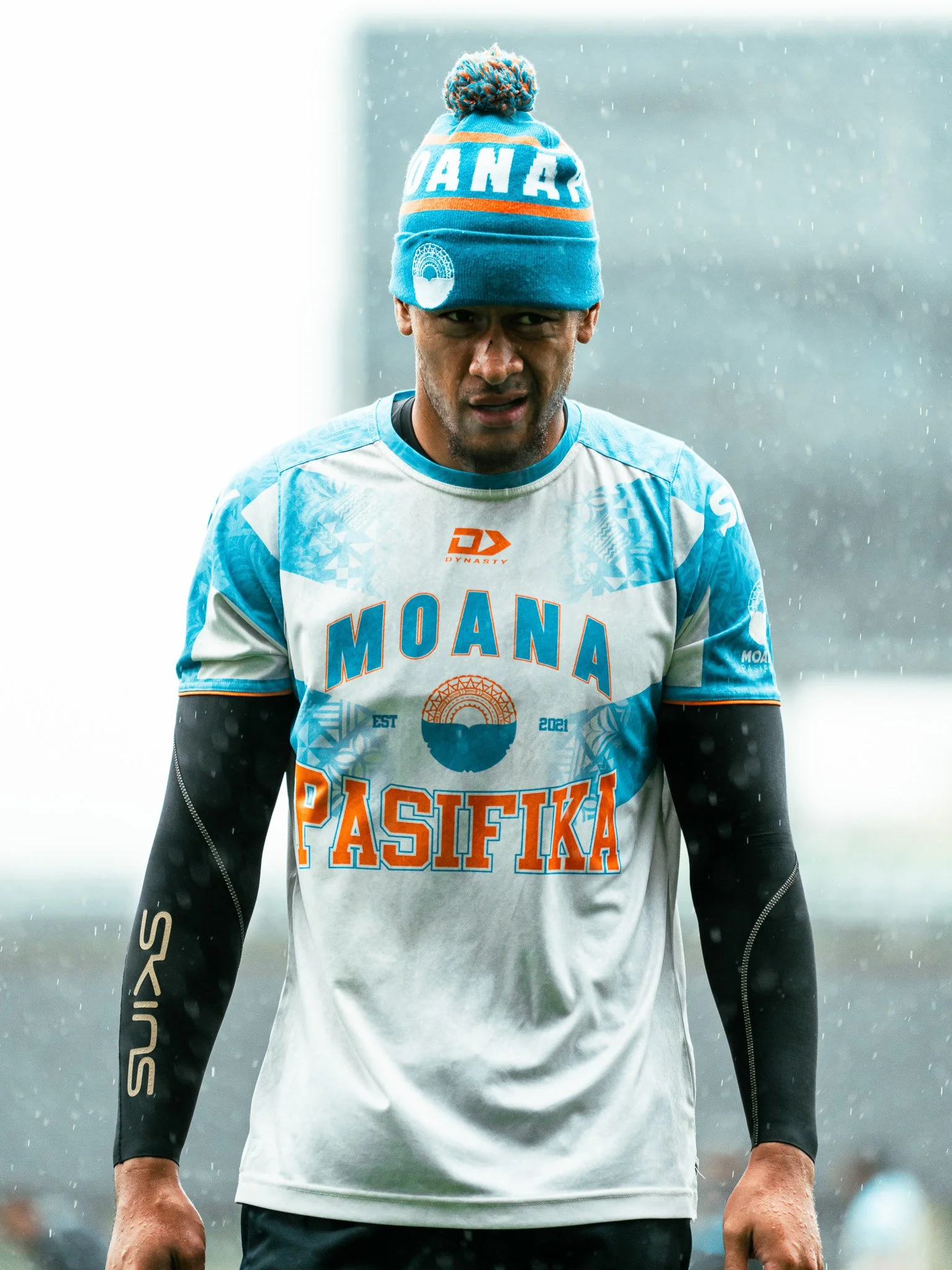 An athlete wearing a blue and white sports uniform with orange accents, a blue beanie with a pom-pom, and black long-sleeve undergarments, standing outdoors in rainy weather.