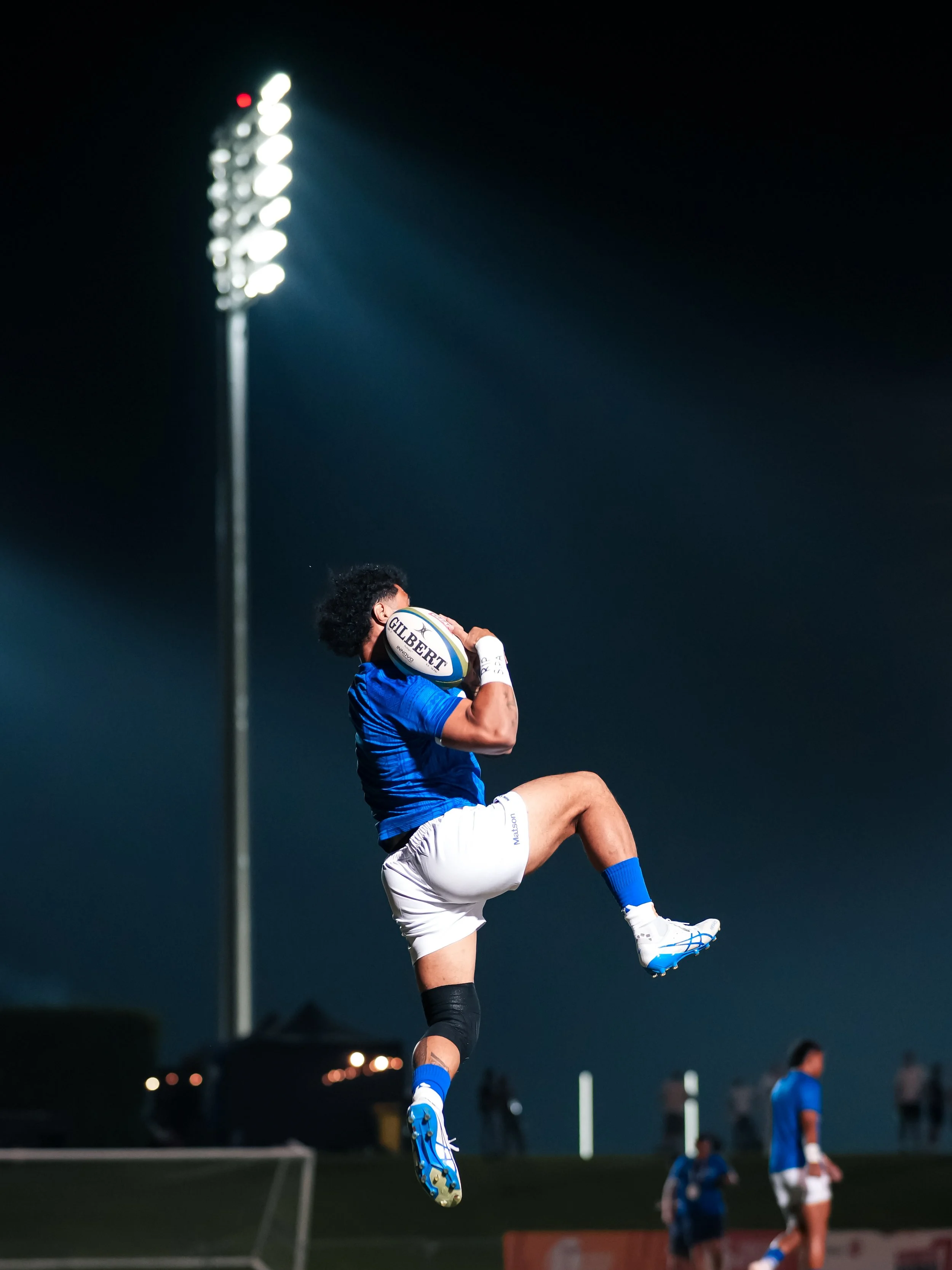 A rugby player in a blue and white uniform leaps to catch a rugby ball during a night game, with a stadium floodlight shining brightly in the background.