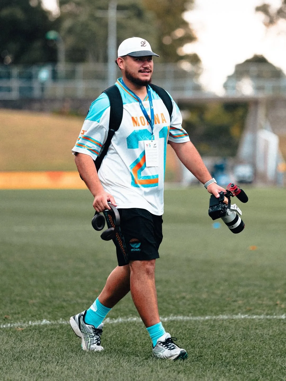 A man dressed in a football jersey, black shorts, teal socks, and sneakers walking across a rugby field, holding a camera and a pair of headpones.