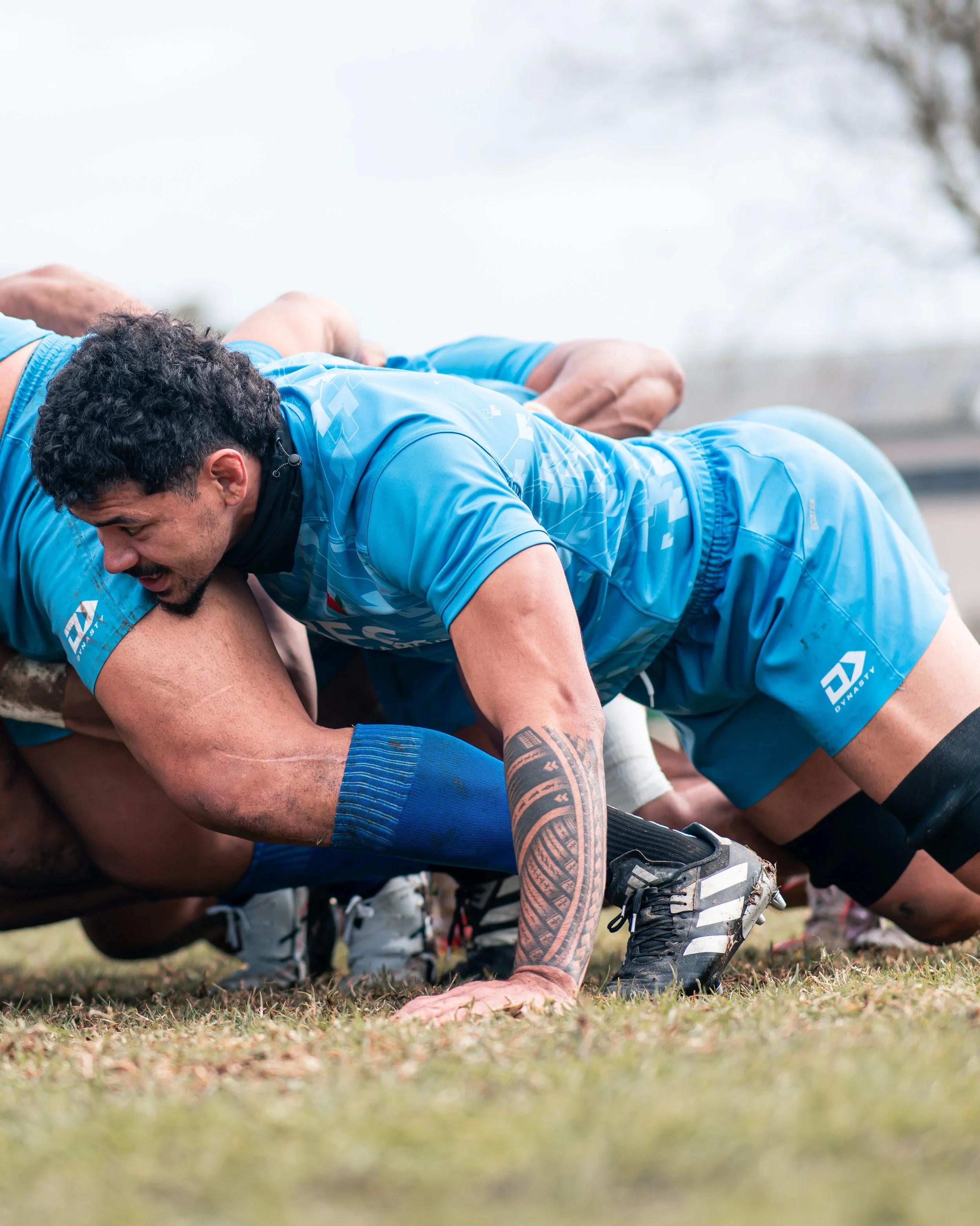 Two rugby players in blue uniforms competing for the ball on the field, with one player on top trying to reach the ball while the other is on the ground.