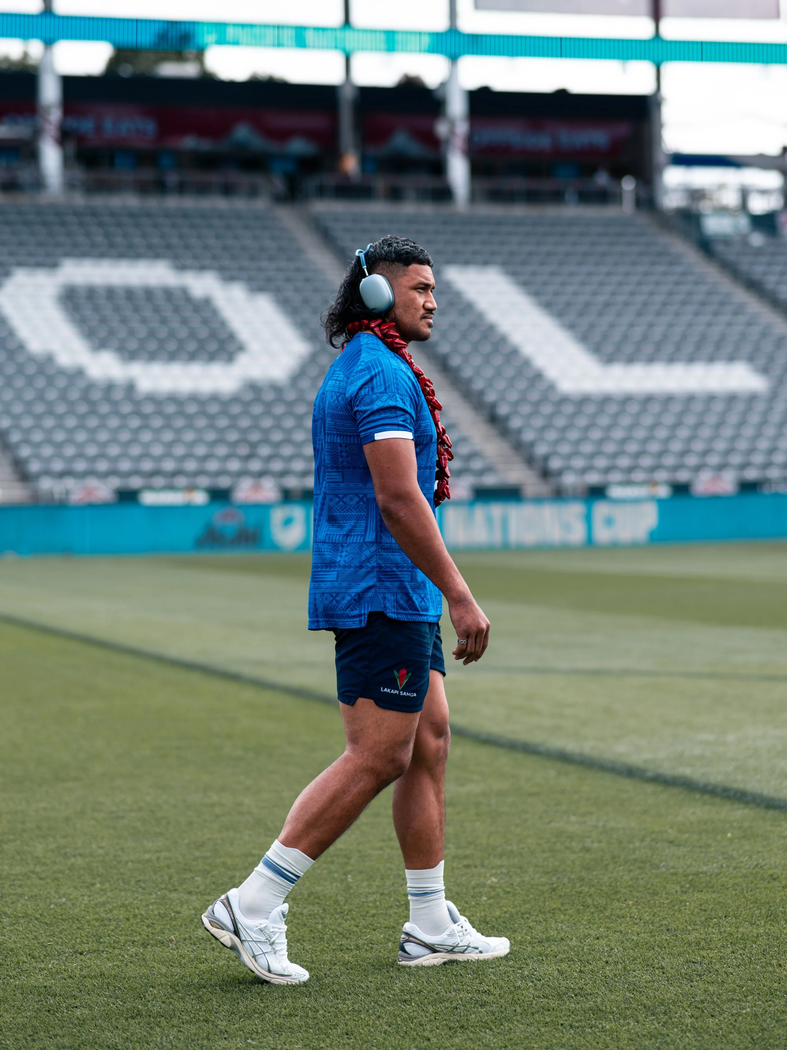 A man wearing a blue sports jersey, navy shorts with a logo that reads 'Lakapai Samoa', white socks with blue stripes, white sneakers, and a red lei is walking on a football field. He is wearing large headphones and has medium-length wavy hair. The s