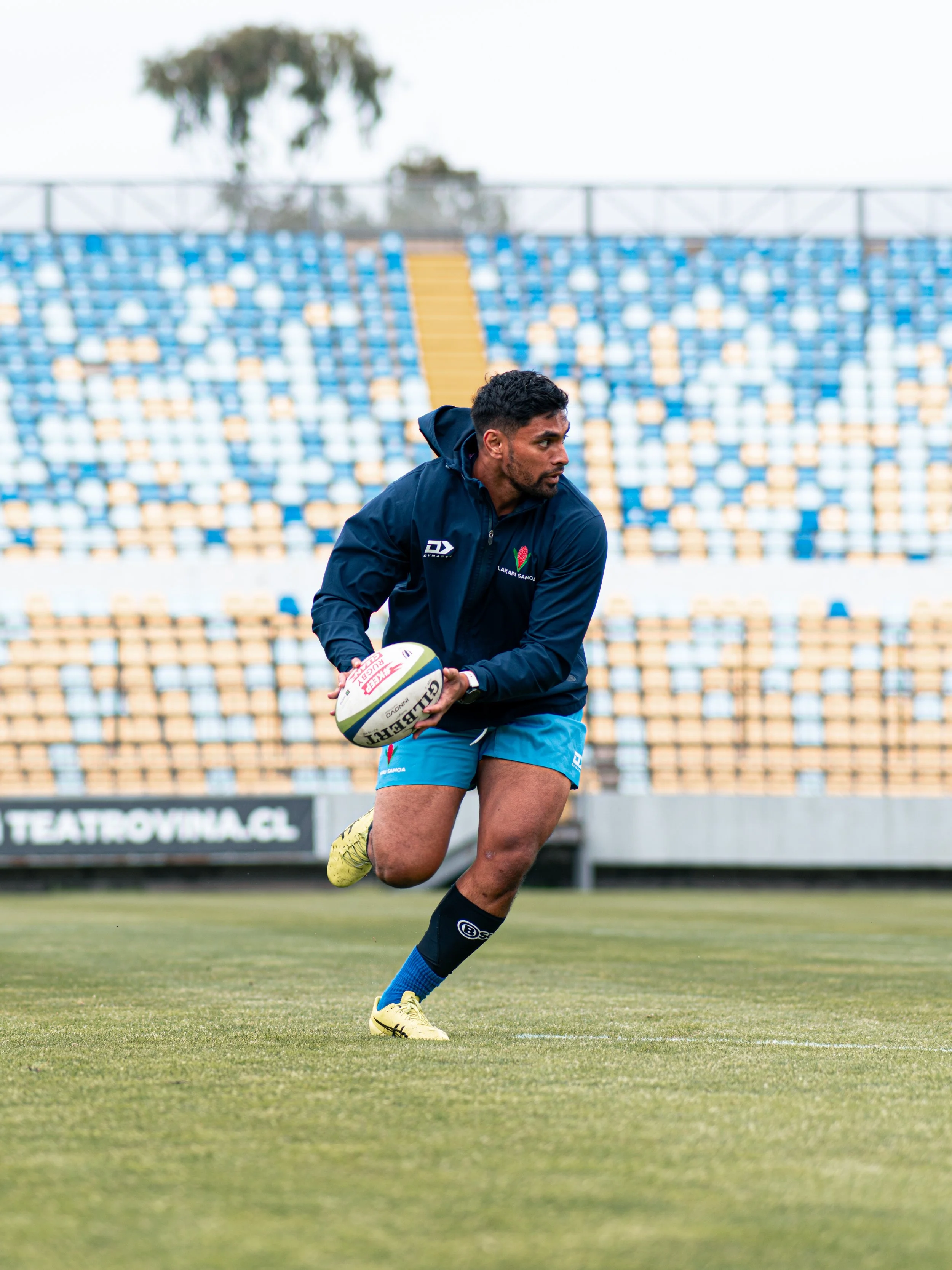 A rugby player in a dark blue jacket and blue shorts running with a rugby ball on a grassy field in a stadium with empty yellow and blue seats.