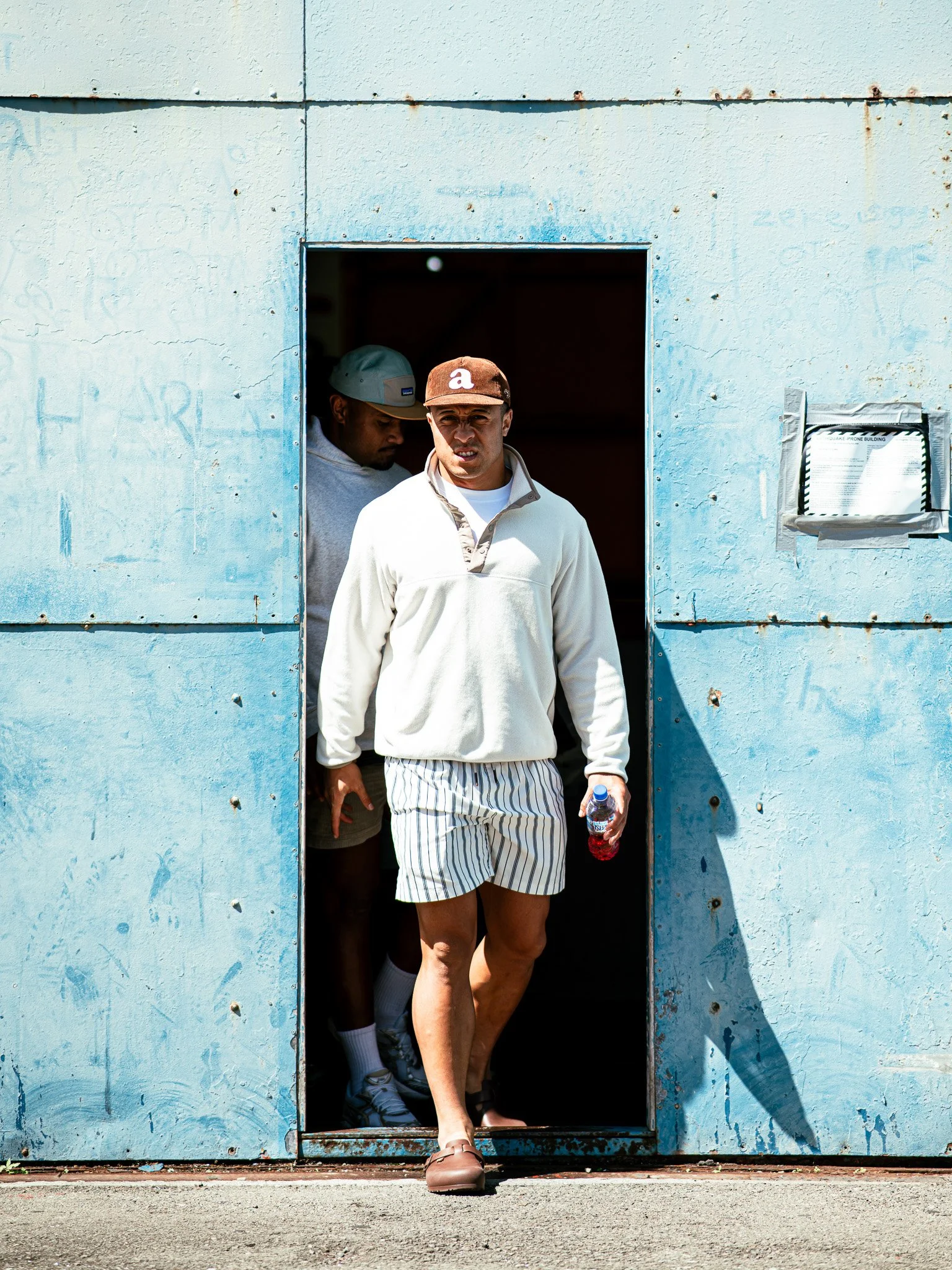 A man walking out of a small tunnel in a weathered light blue building, holding a water bottle, dressed in casual sportswear including a brown cap, white sweatshirt, striped shorts, and sandals, with another person partially visible behind him.