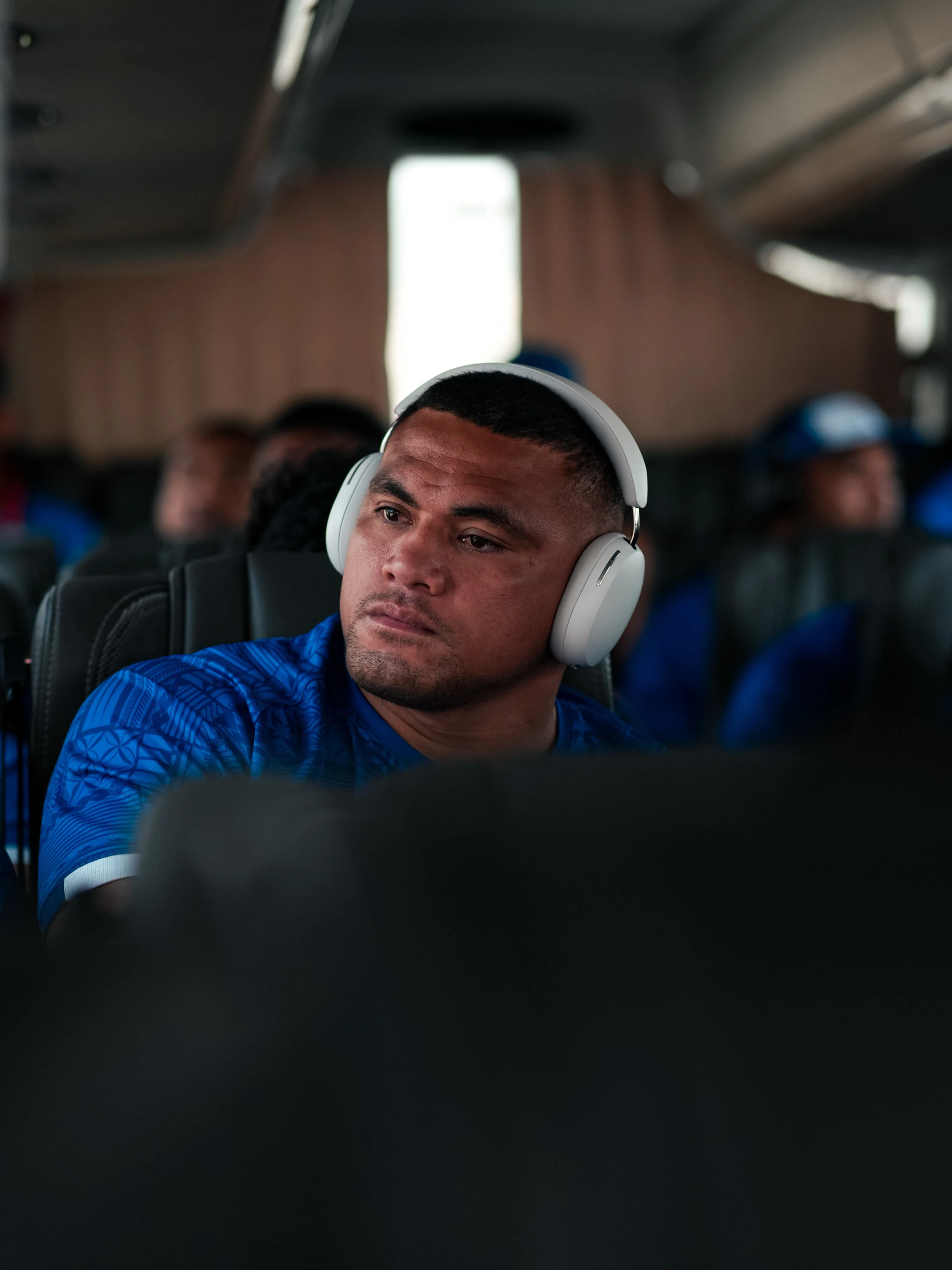 A young man with short dark hair and a beard, wearing a blue sports jersey and white over-ear headphones, sitting on a bus or airplane, looking thoughtfully to the side. Other passengers are visible in the background.