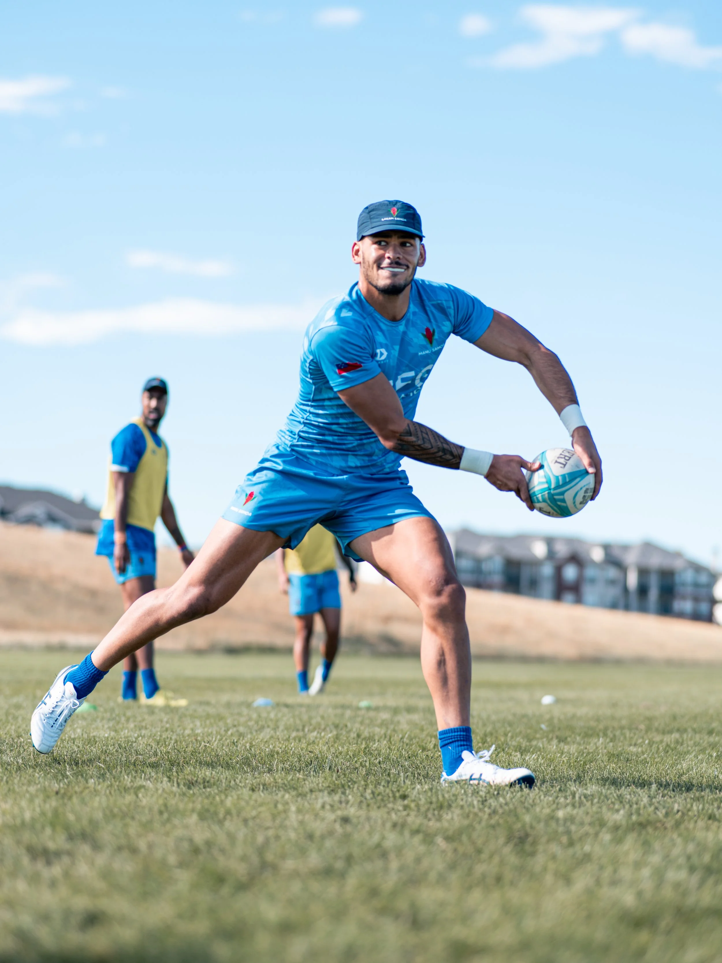 A man in blue sports uniform with a cap is playing rugby and holding a rugby ball on a grassy field with a blue sky and few clouds, with other players and residential houses in the background.