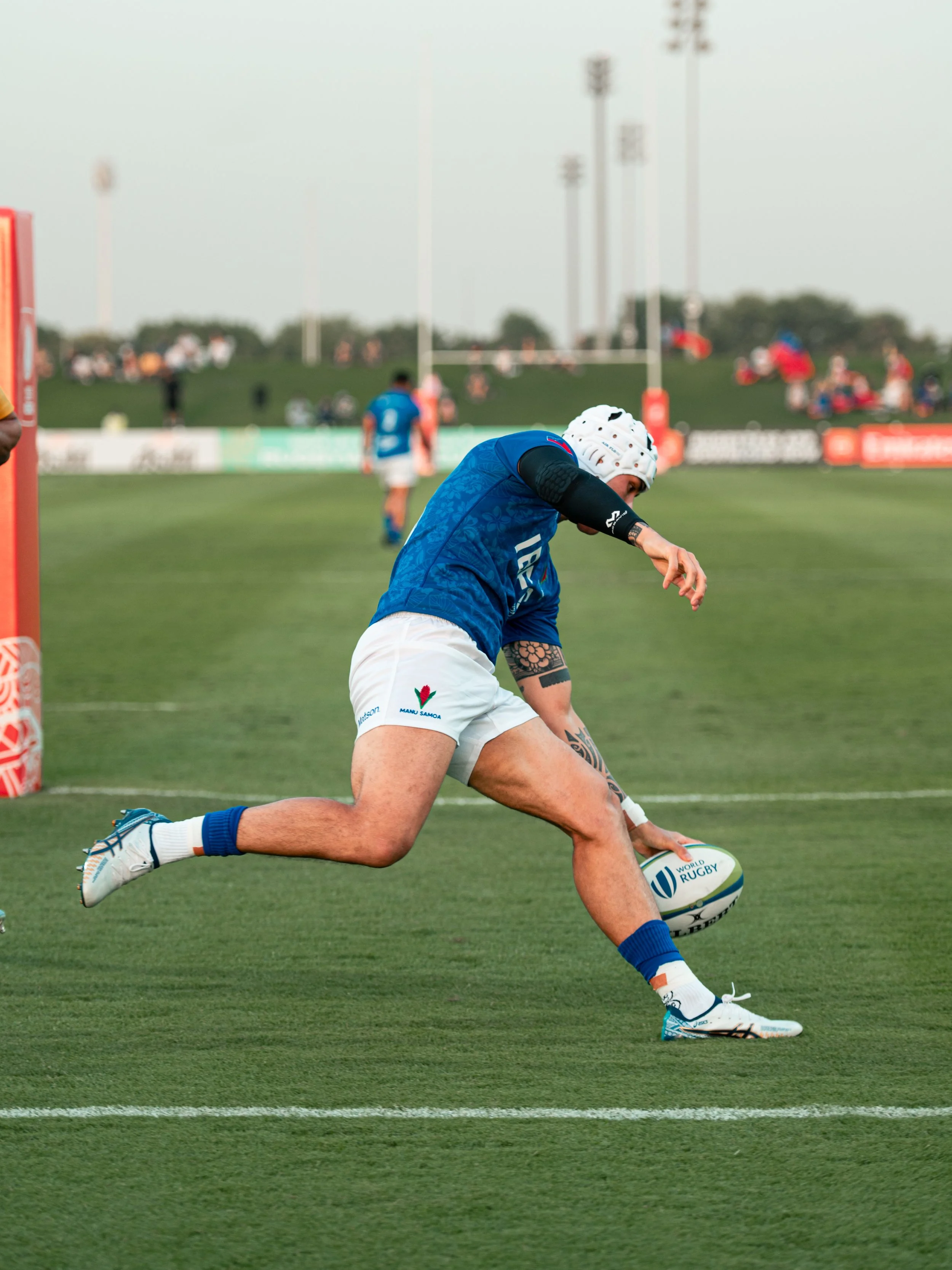 A rugby player in a blue jersey and white shorts, wearing a helmet and sporting tattoos, is diving to score a try on a rugby field, with spectators in the background.